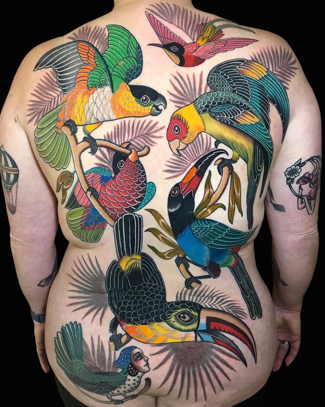 Colorful back tattoo featuring multiple parrots and tropical birds perched on branches with spread wings in vibrant greens, yellows, reds, and blues.