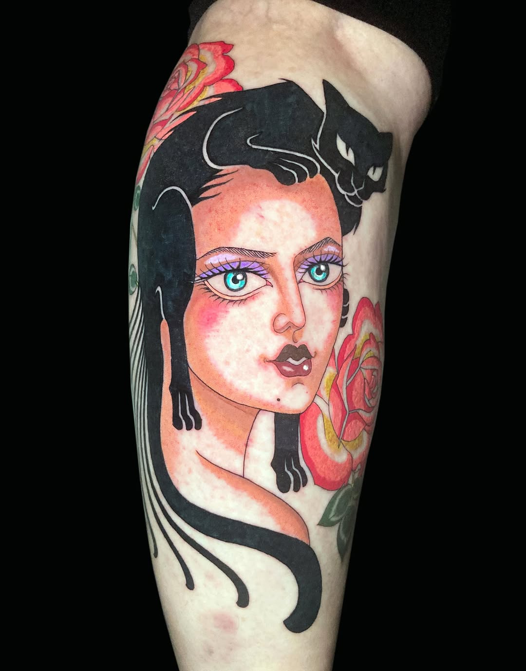 Colorful tattoo on a leg featuring a woman's face with turquoise eyes, black cats on her shoulders, and red flowers beside her head.