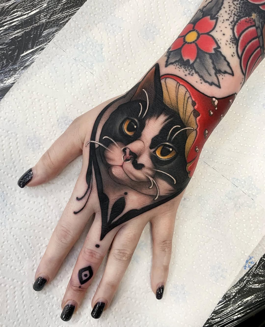 Black cat face with golden eyes and white whiskers on hand, with red flowers and leaf designs extending up the forearm.