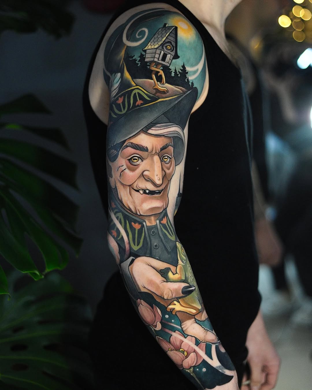 Full-sleeve arm tattoo featuring a smiling gnome character with a pointed hat above a whimsical house scene, rendered in multicolored tones on dark background.