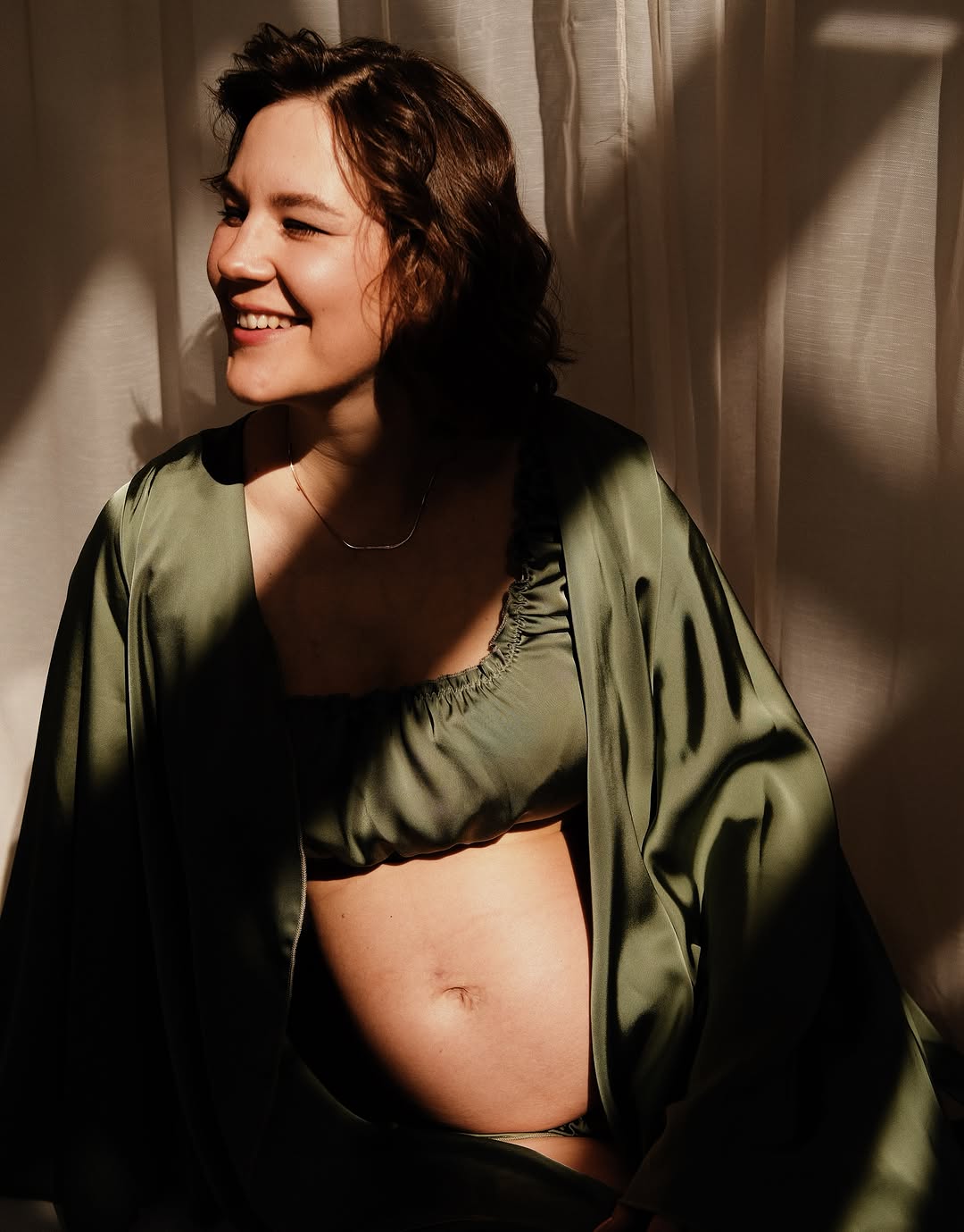 Pregnant woman displaying a small dark tattoo on her abdomen, wearing an olive green open shirt against a neutral background with natural sunlight.