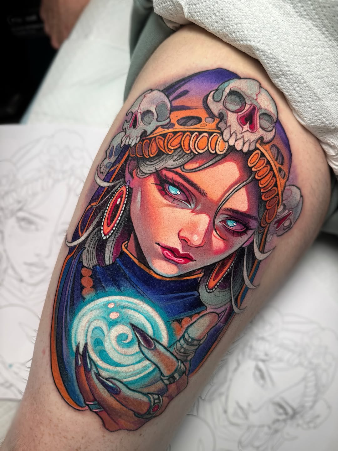 Colorful shoulder tattoo featuring a woman's face with skull crown, blue eyes, red lips, ornate earrings, and a spiral symbol on her chest against a gradient background.