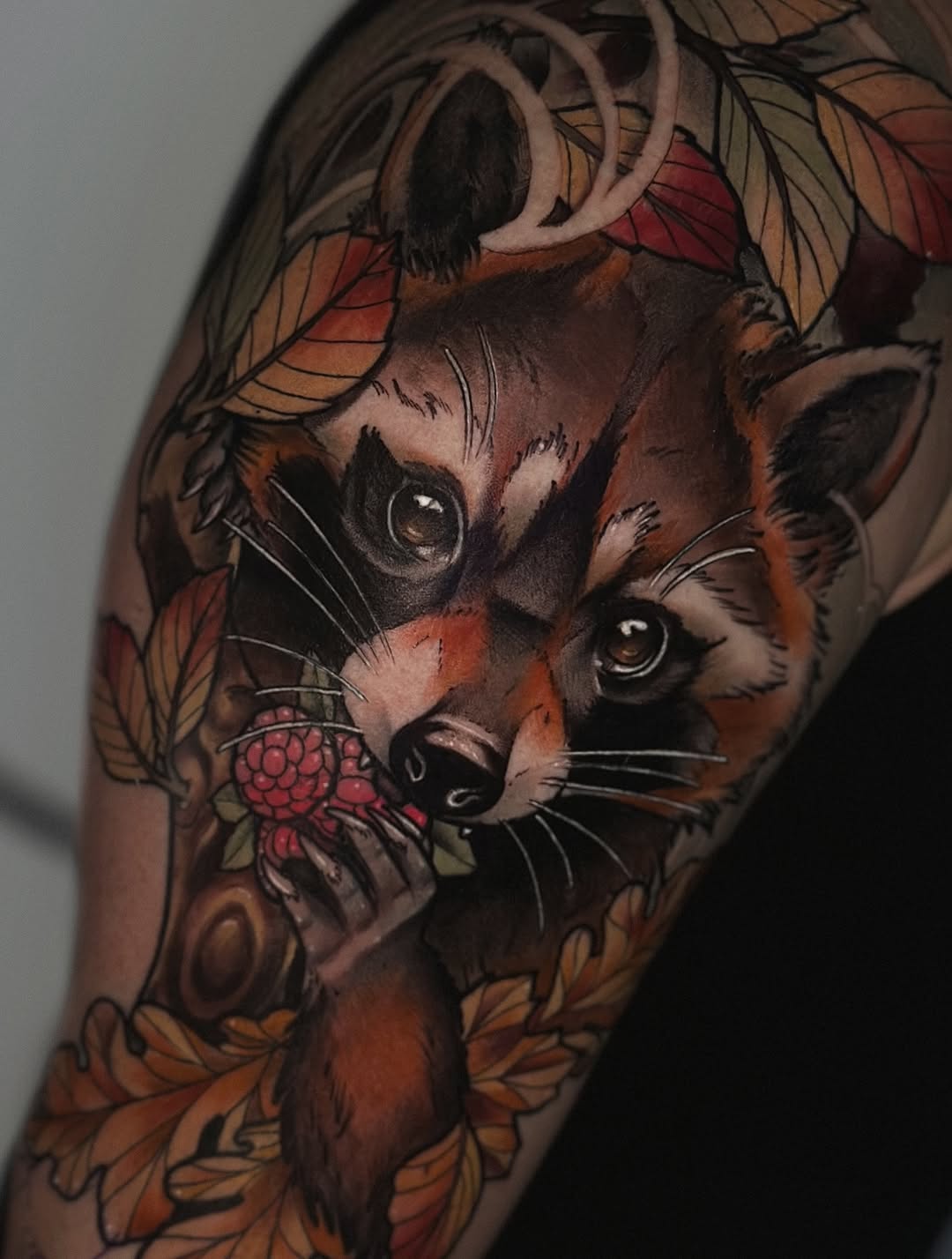 Detailed color tattoo on upper arm featuring two raccoons with autumn leaves and a red raspberry, rendered in brown, orange, red, and black tones.