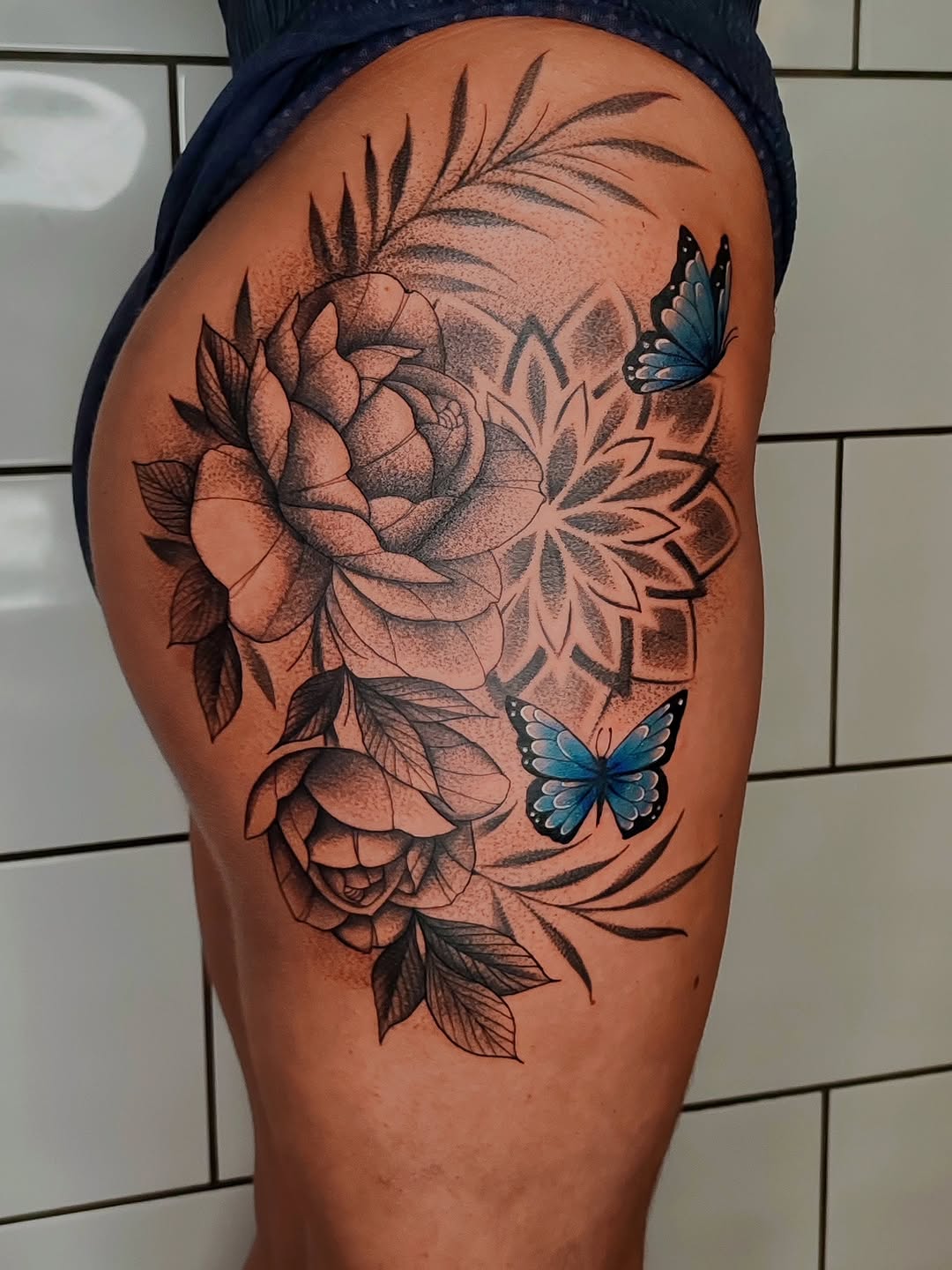 Floral tattoo on thigh featuring roses and decorative leaves in black and brown with two blue butterflies.