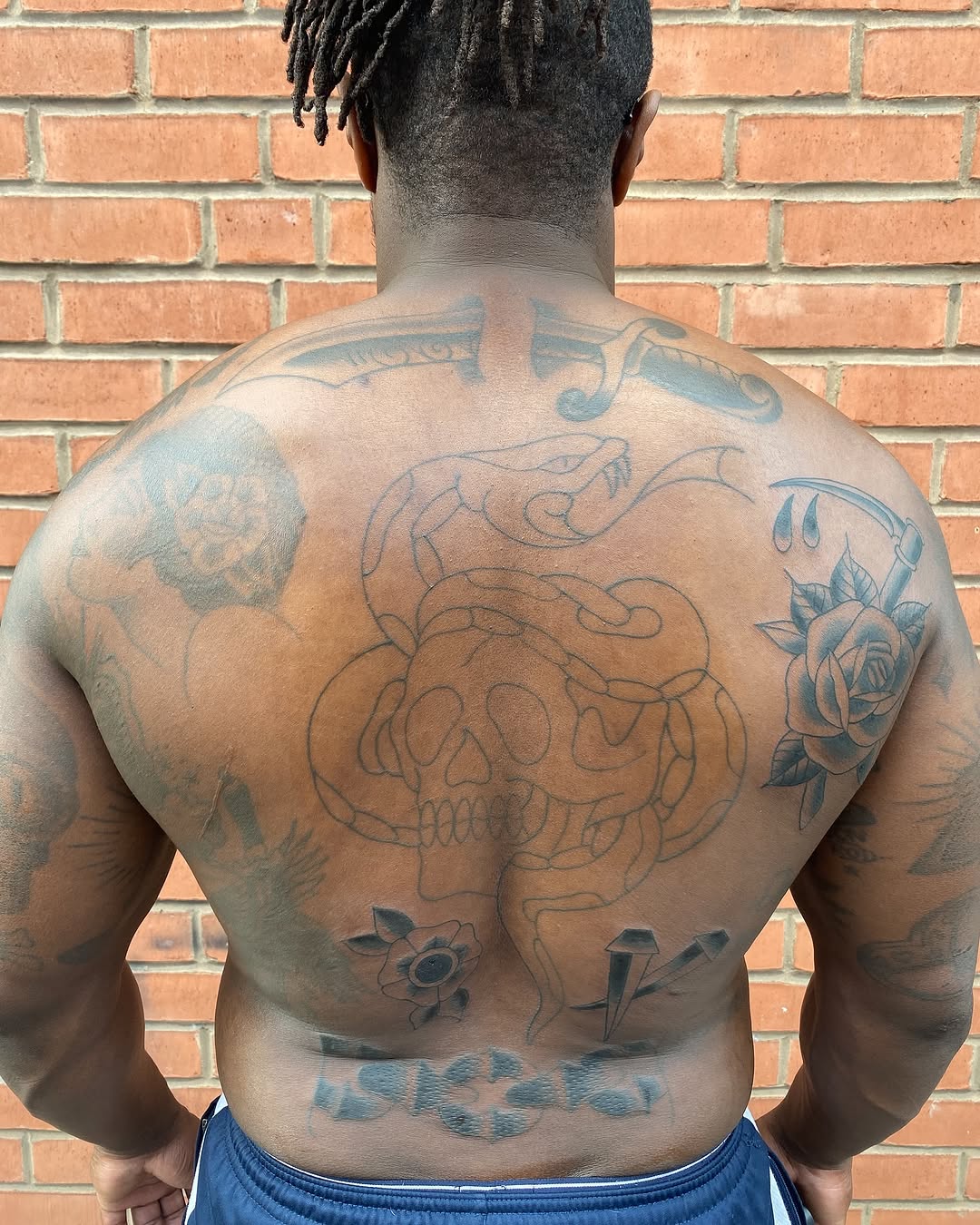 Rear view of a shirtless person displaying a large skull with serpents on the back, flanked by roses and additional small tattoos on the arms and lower back.