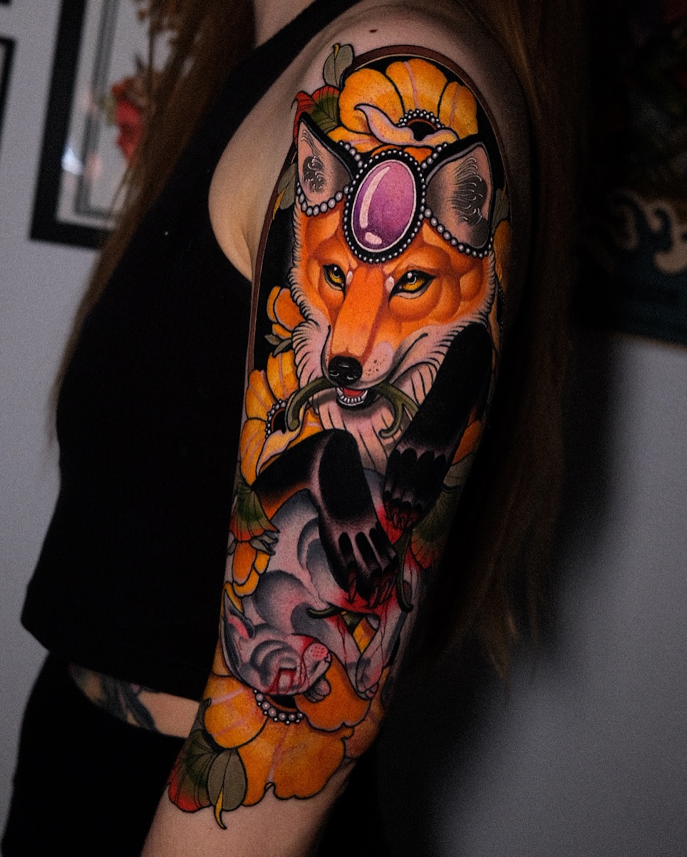 Colorful fox head tattoo with ornamental headpiece on upper arm, featuring orange flowers, purple gemstone, and skull motif below in vibrant hues.