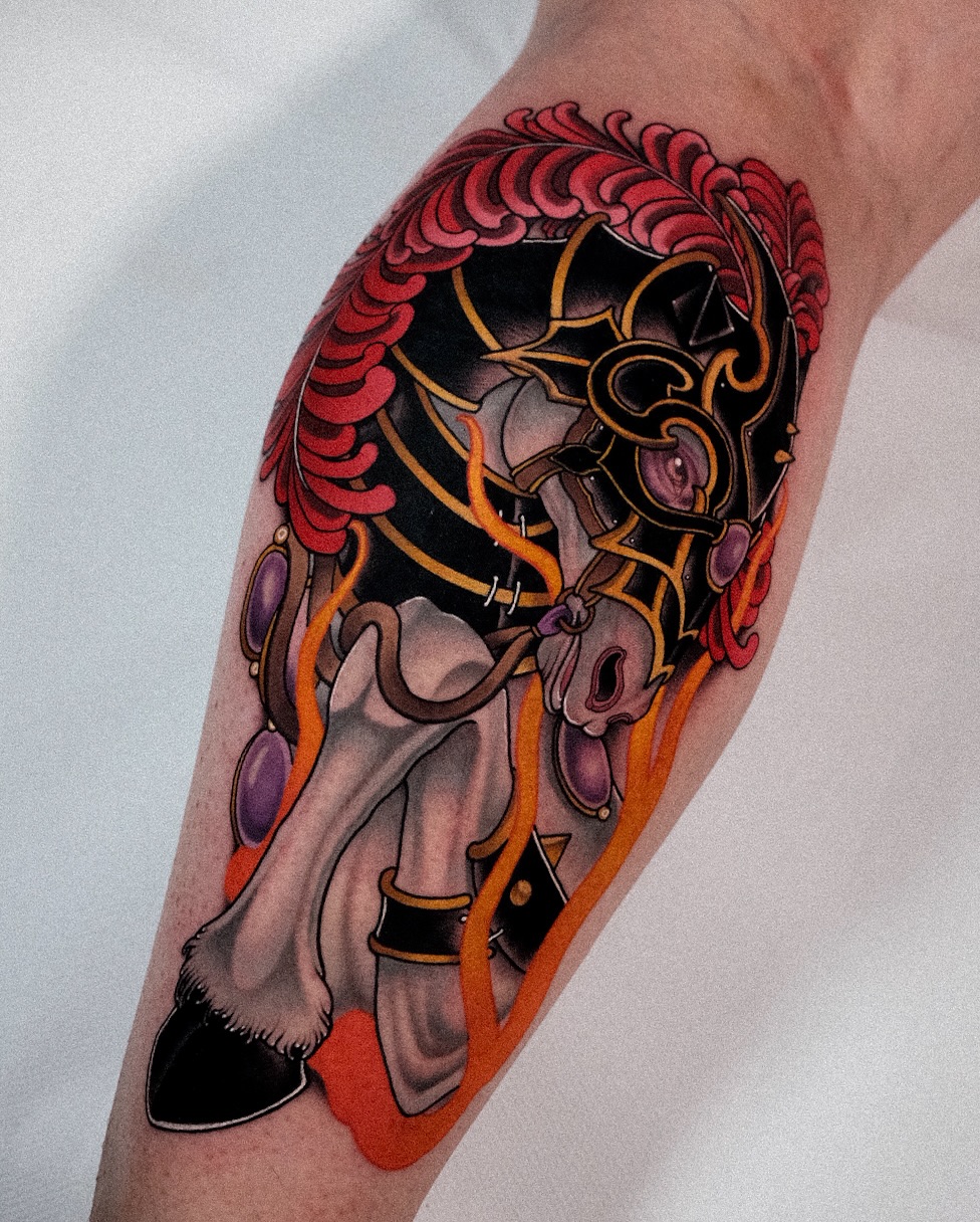 Colorful tattoo on a calf featuring a mythical horned creature with red scaled wings, orange and gold accents, and purple gemstone details against black shading.