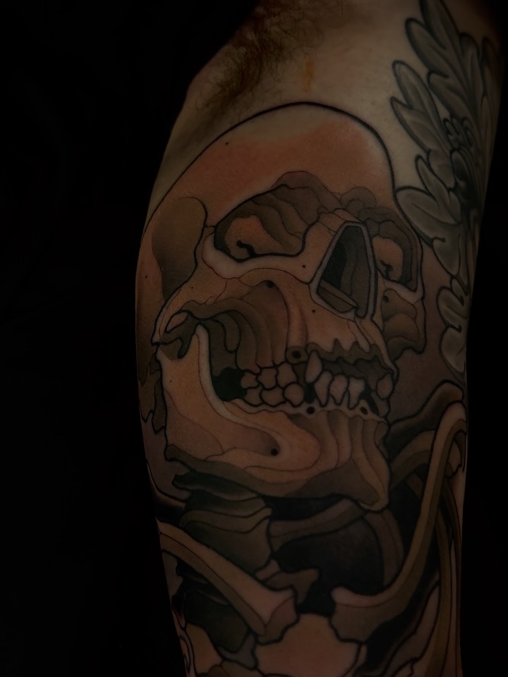 Skull tattoo on upper arm with shading and line work, featuring anatomical bone details and decorative elements on the shoulder.