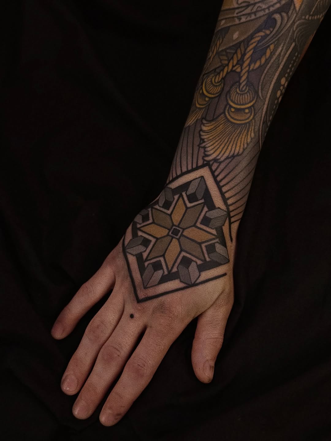 Detailed geometric and ornamental tattoo on forearm and hand featuring a starred mandala pattern and decorative upper arm work in brown and black ink.