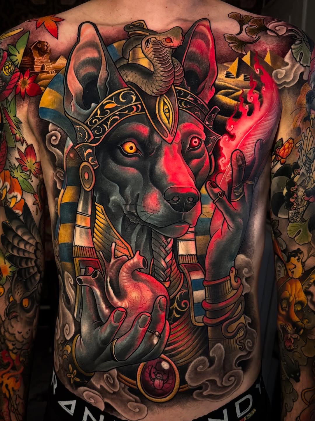 Large-scale back tattoo featuring an Egyptian jackal deity head with golden ornaments, surrounded by colorful flowers, pyramids, and decorative elements in red, gold, and black tones.
