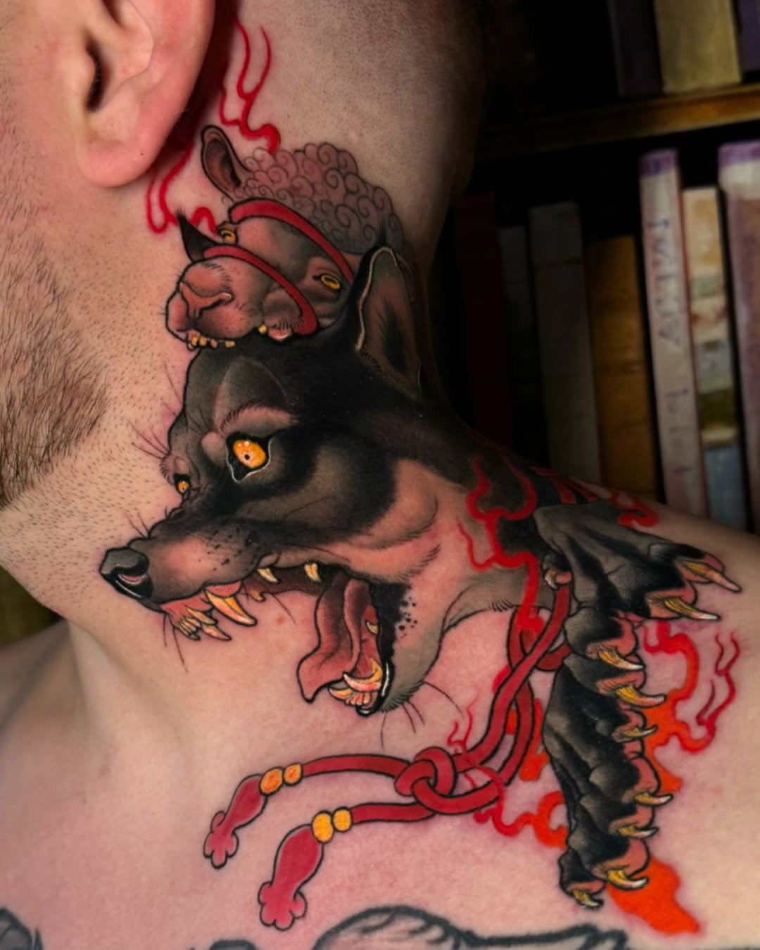 Colorful neck tattoo featuring three stacked demons or creatures with golden eyes, red flames, and ornamental details in red, black, and orange.
