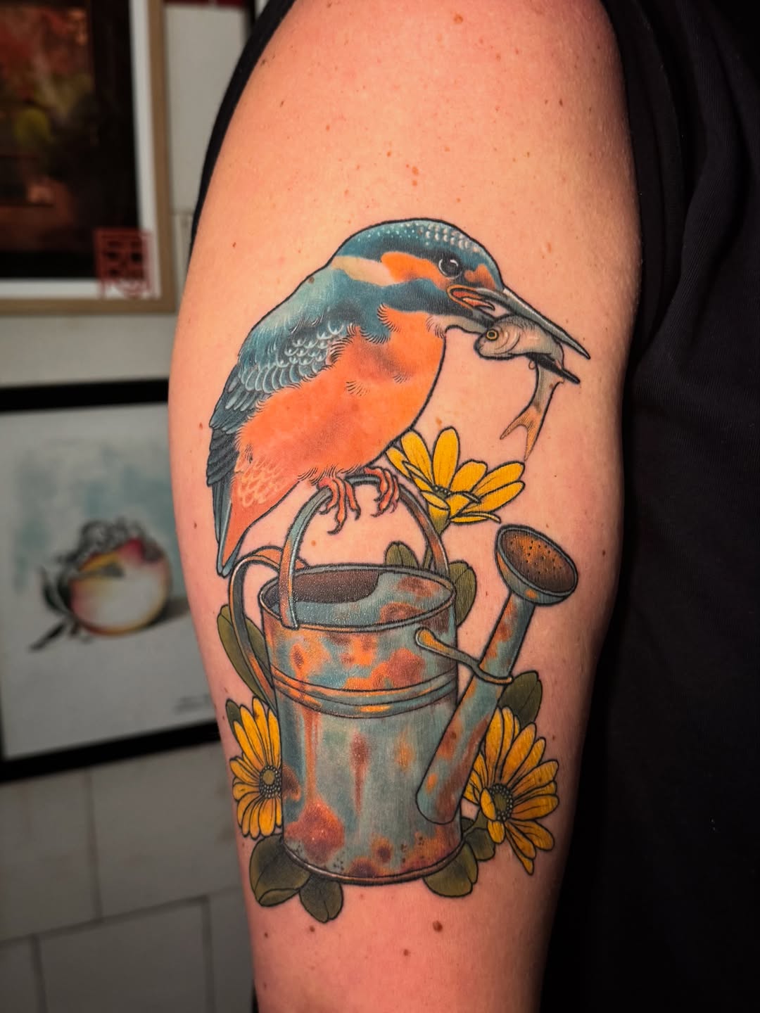 Colorful kingfisher tattoo perched on a watering can with yellow flowers, displayed on the upper arm in orange, teal, and gold tones.