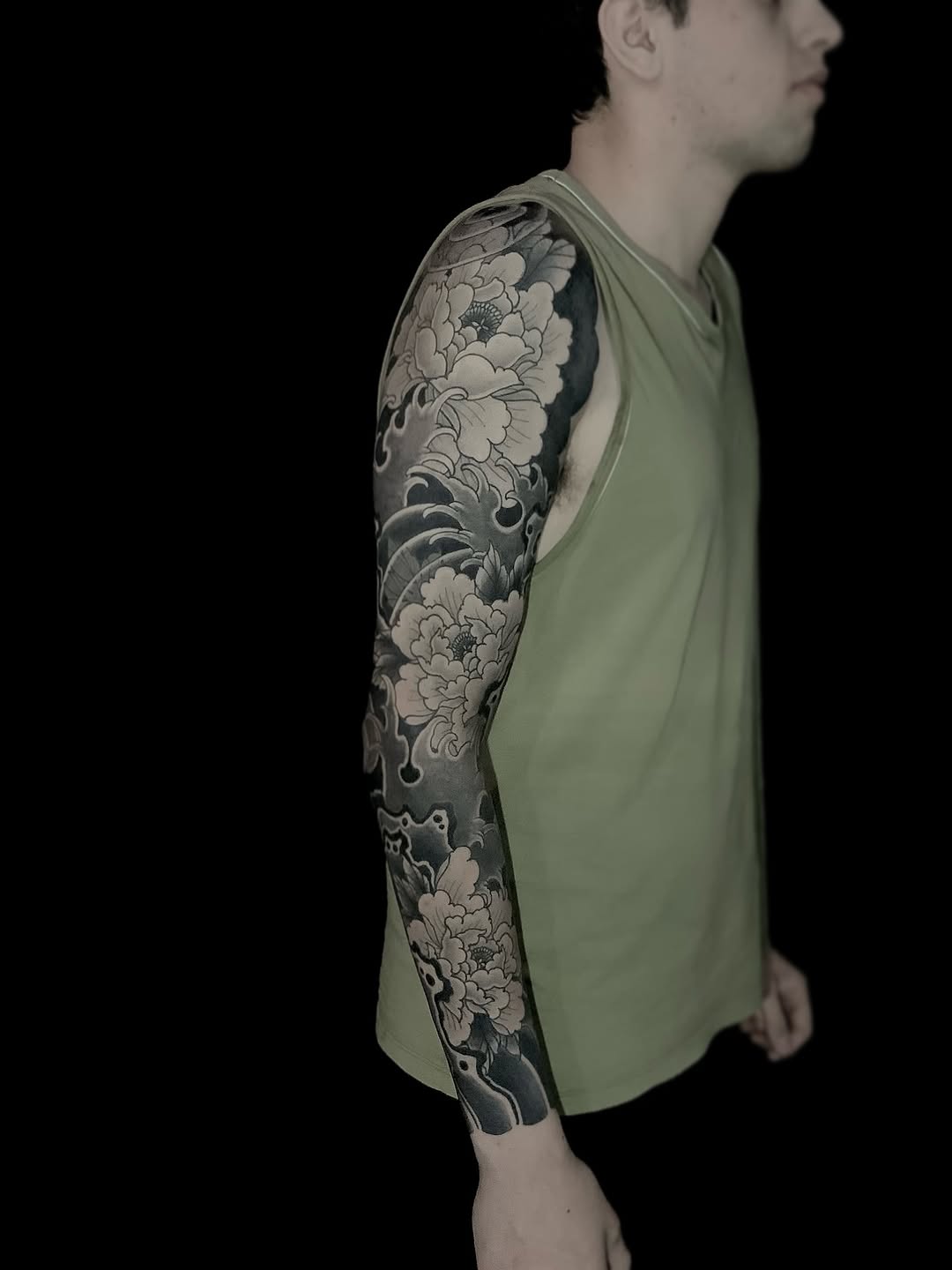 Man wearing a light green sleeveless shirt displaying a full arm sleeve tattoo featuring large detailed flowers and ornamental scrollwork in black and gray ink.