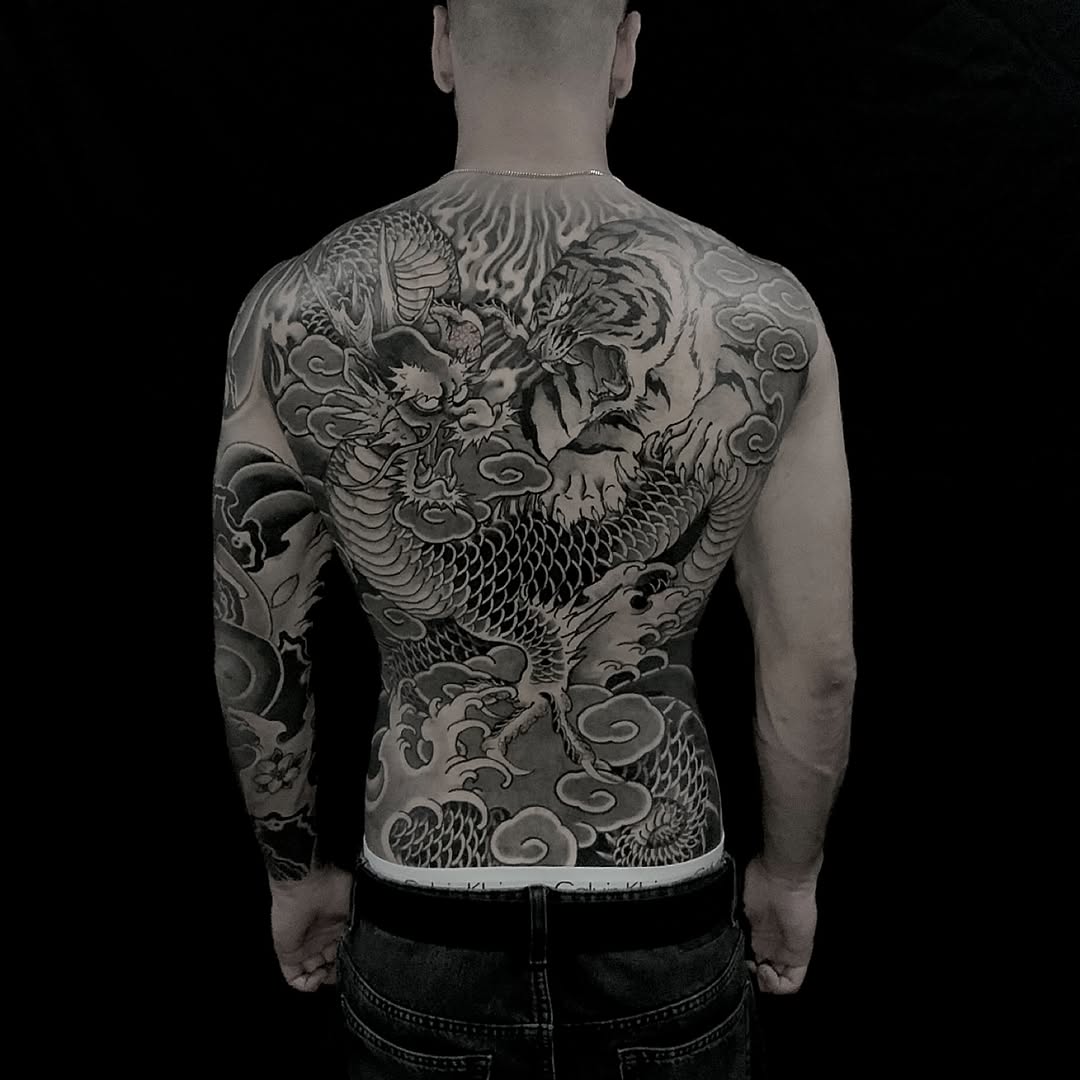 Rear view of a shirtless man displaying a full back tattoo featuring dragons, clouds, and ornamental scrollwork in grayscale ink.