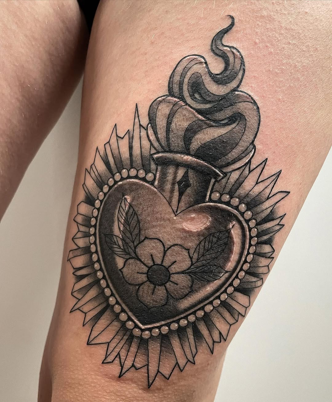 Ornate heart tattoo with flower center, flame crown, pearl border, and spiky wing details in black and gray ink on upper arm.