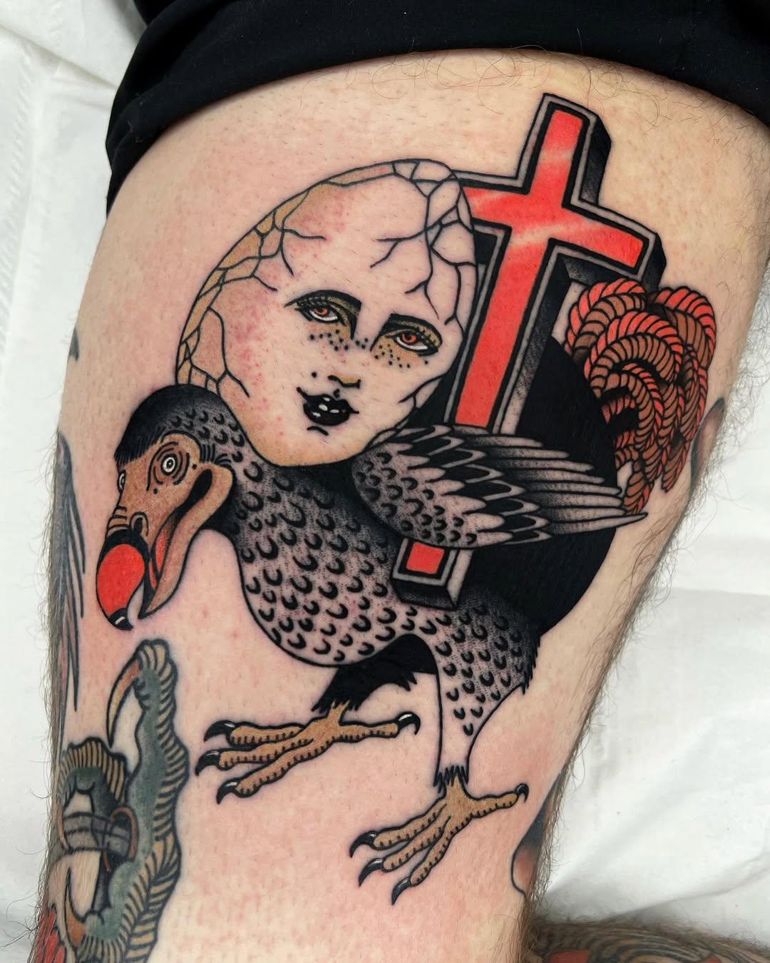 Colorful tattoo on thigh featuring a pale-faced angel with wings and a red cross, accompanied by a bird and decorative elements in red, black, and tan.