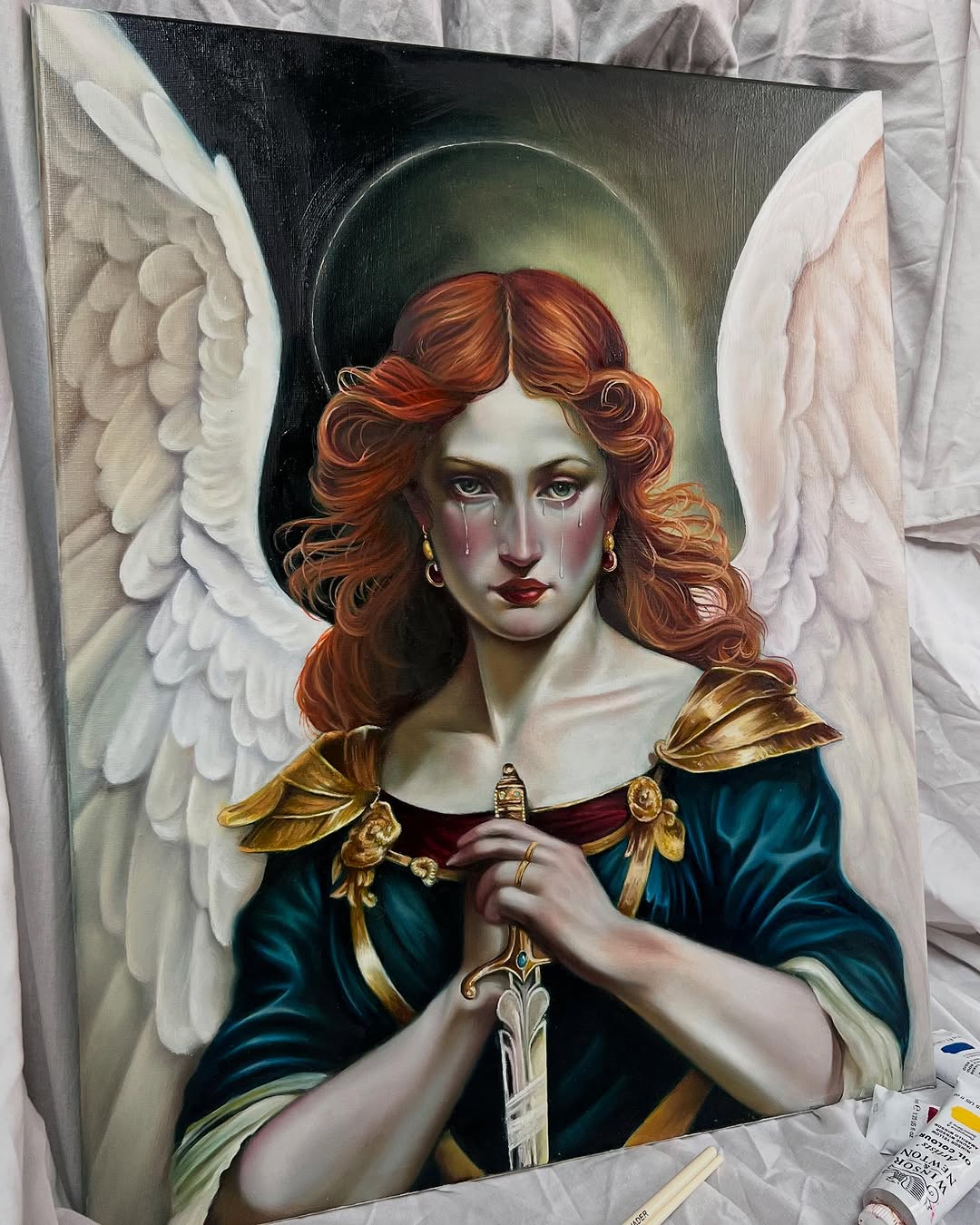Portrait painting of an angel with copper-red wavy hair, pale complexion, tears on cheeks, wearing dark teal robe with gold shoulder armor and holding a ornate sword.