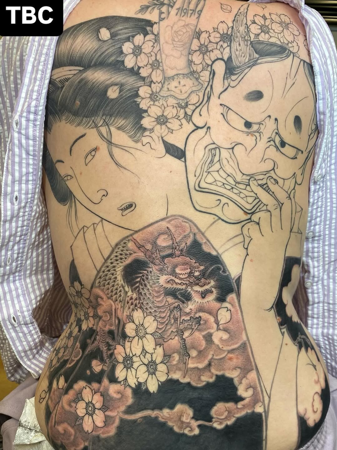 Back tattoo featuring two geisha faces with flowers in their hair, a dragon with clouds and floral elements in sepia and black ink.