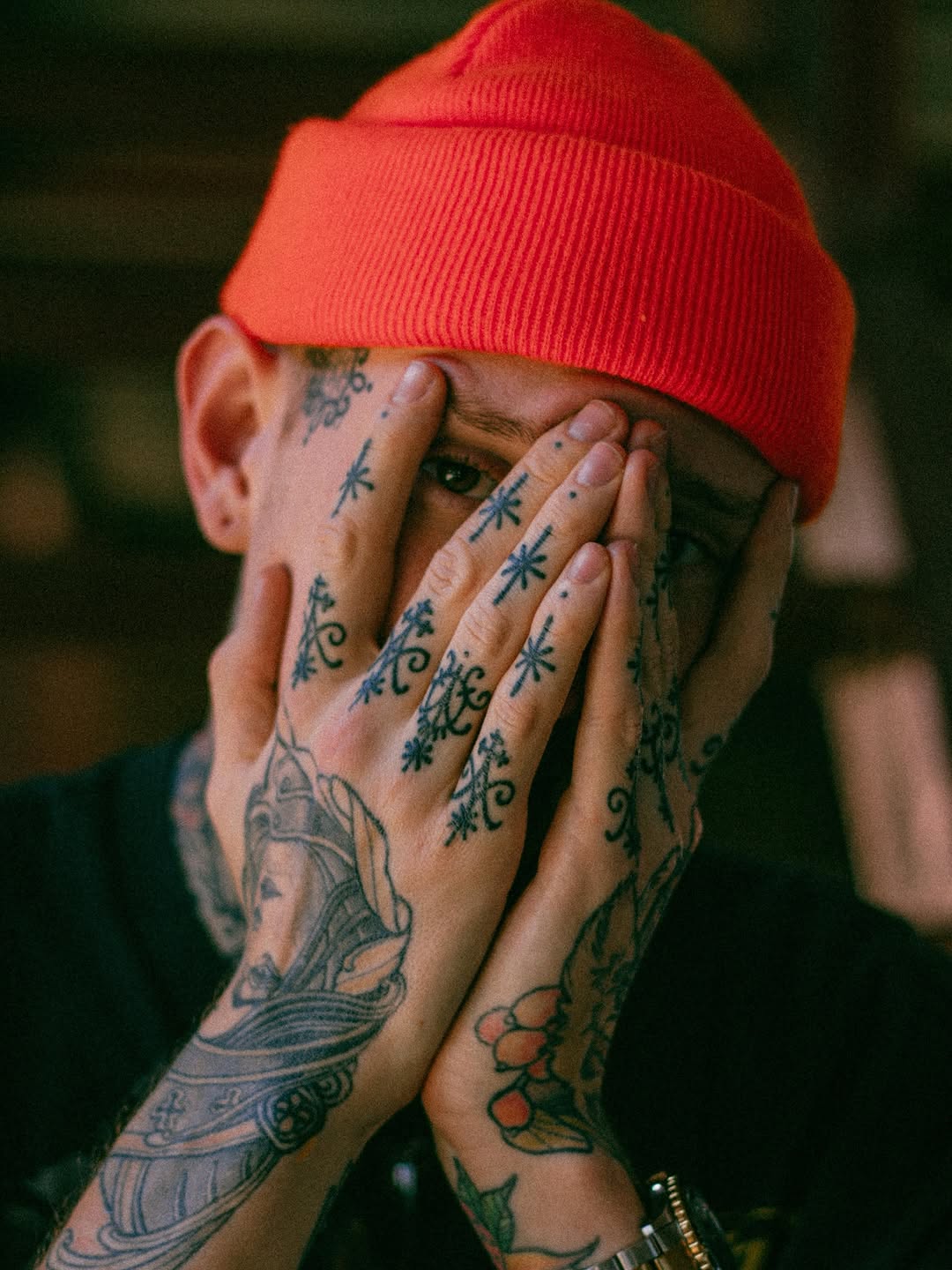 Person wearing a red beanie with heavily tattooed hands and arms featuring botanical designs, geometric patterns, and ornamental details in black and green ink.