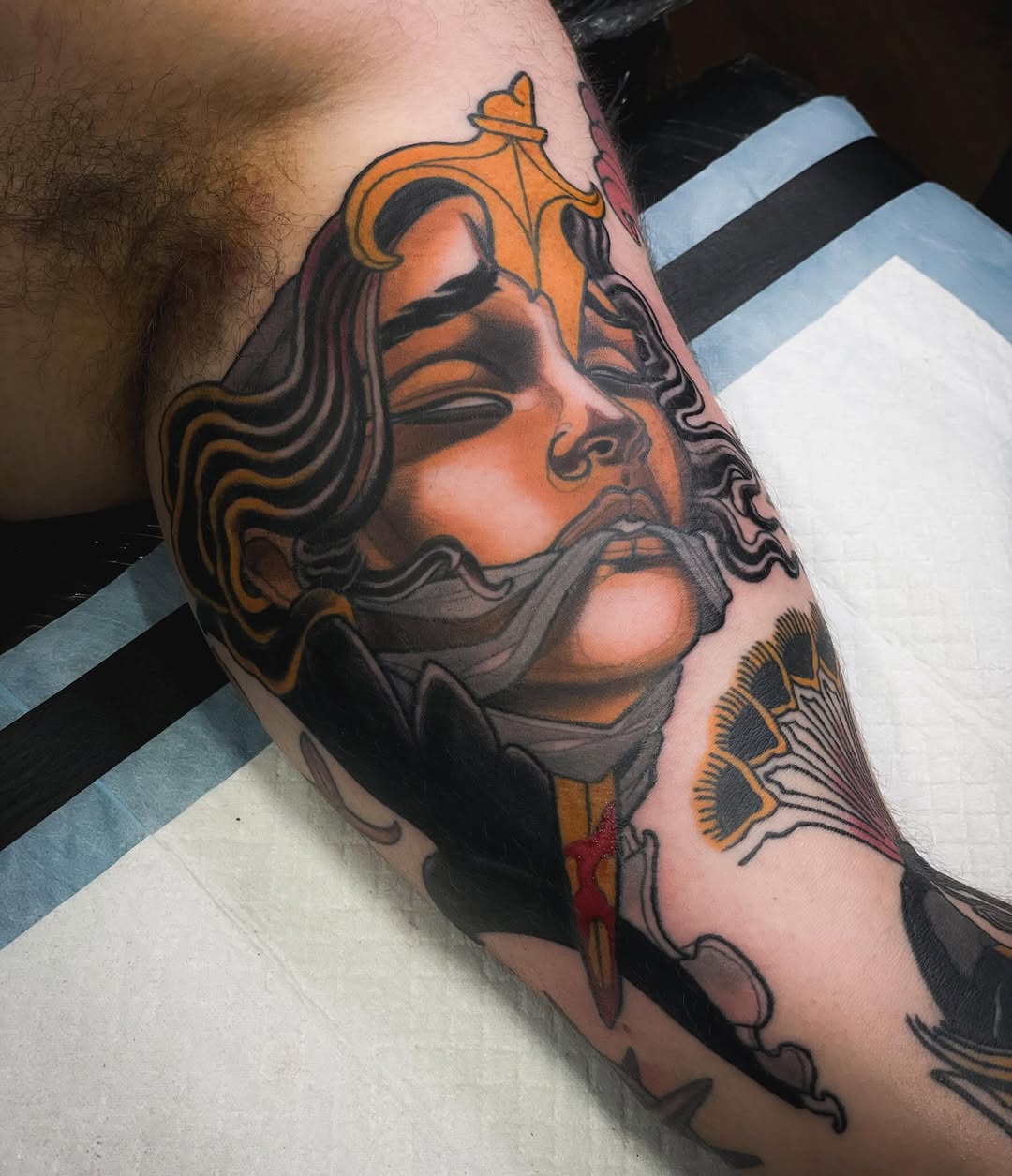 Colorful portrait tattoo of a bearded male face with orange and gold accents, decorative elements, and wing designs on the forearm.