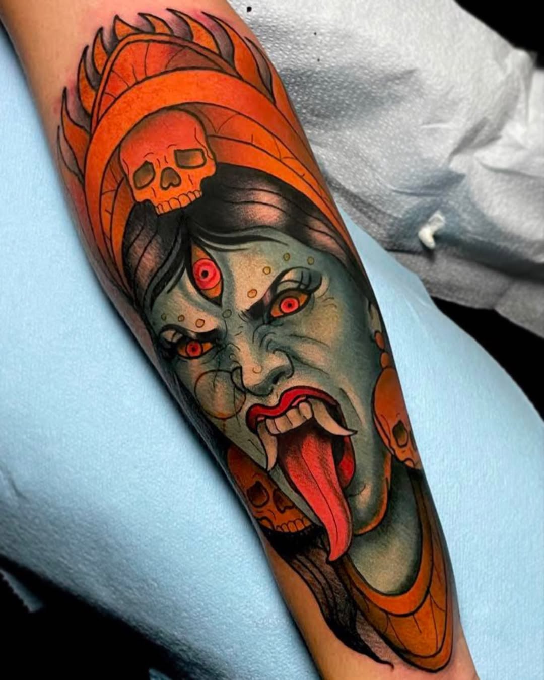 Colorful arm tattoo featuring a demon face with red eyes and extended tongue, topped with a flaming skull and orange headwrap.