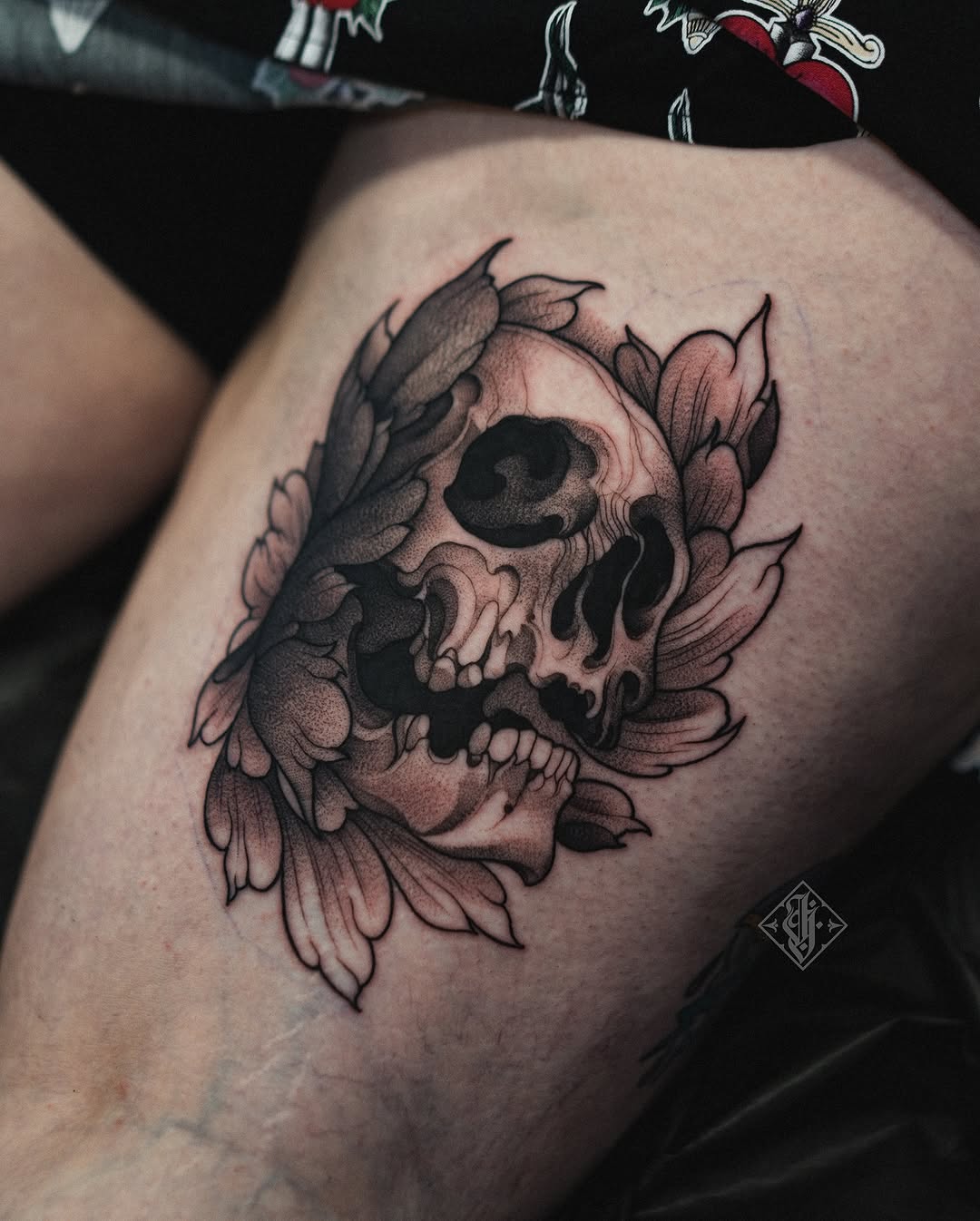 Skull surrounded by floral petals in black and pink ink on the thigh, with fine shading and detail throughout the design.