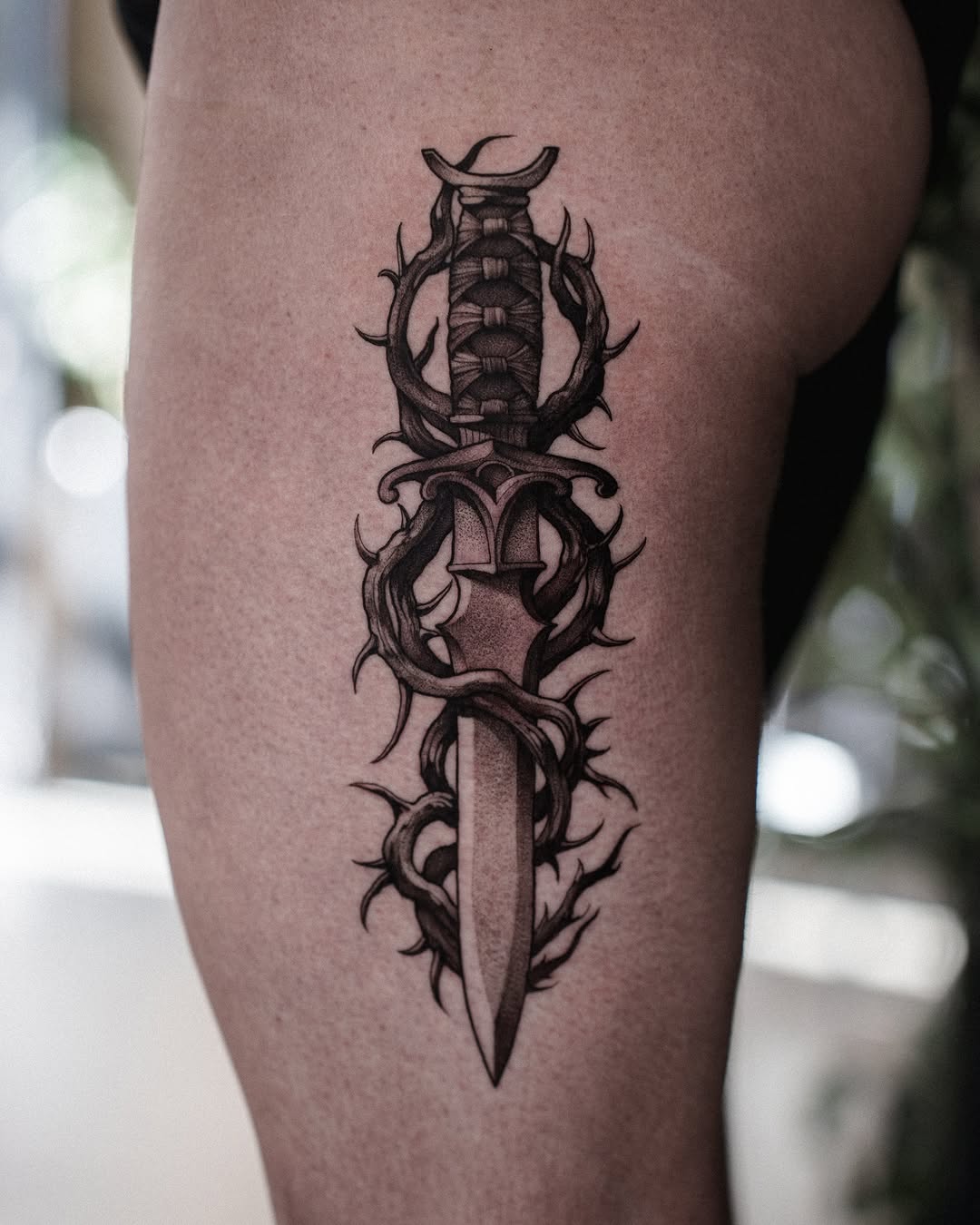 Vertical ornate sword tattoo with thorned vines wrapping around the blade and hilt, featuring a crowned pommel, displayed on the thigh in black ink.
