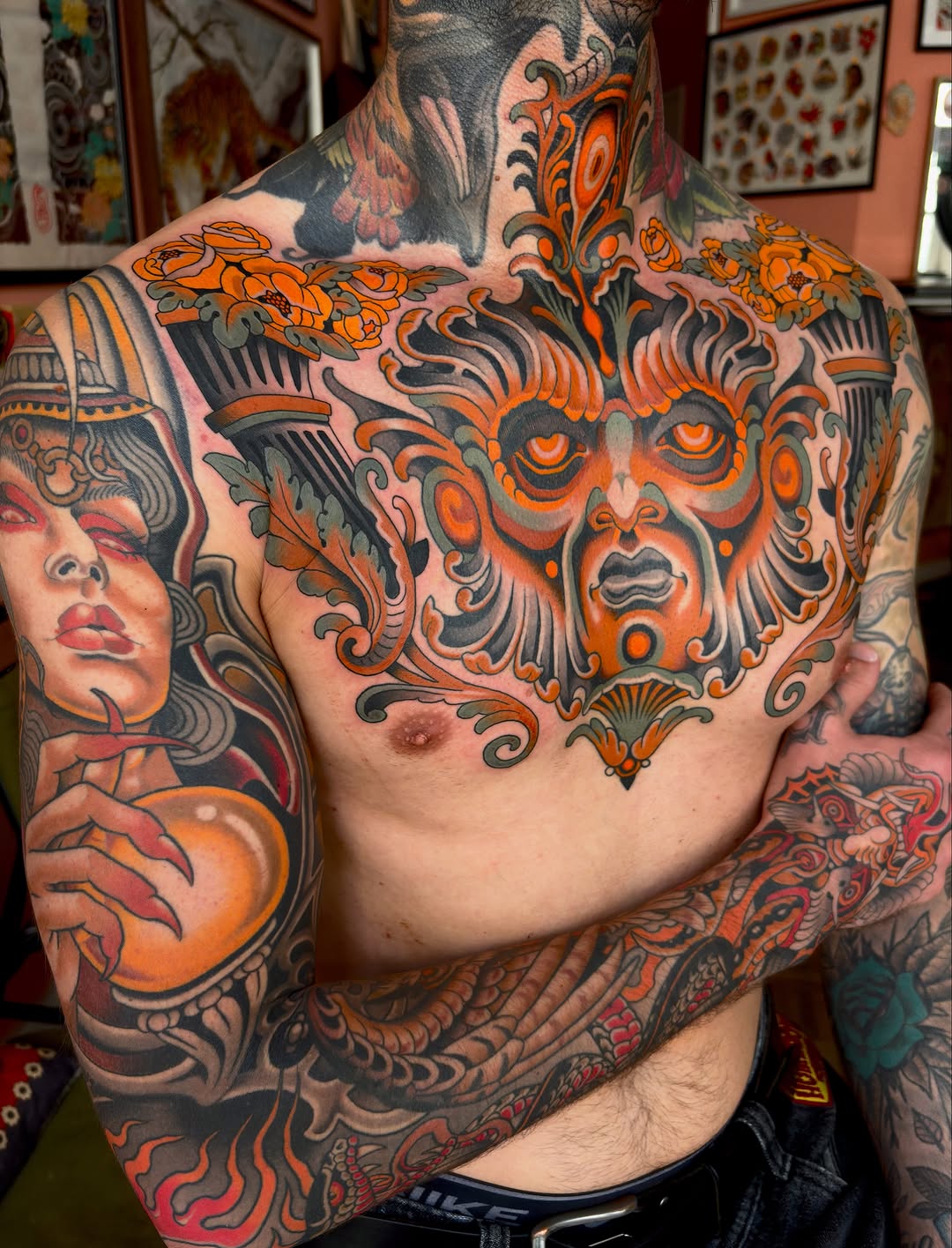 Heavily tattooed male chest and shoulders featuring an ornate orange and black demon face centerpiece with decorative scrollwork, surrounded by additional imagery in orange, red, and black tones.