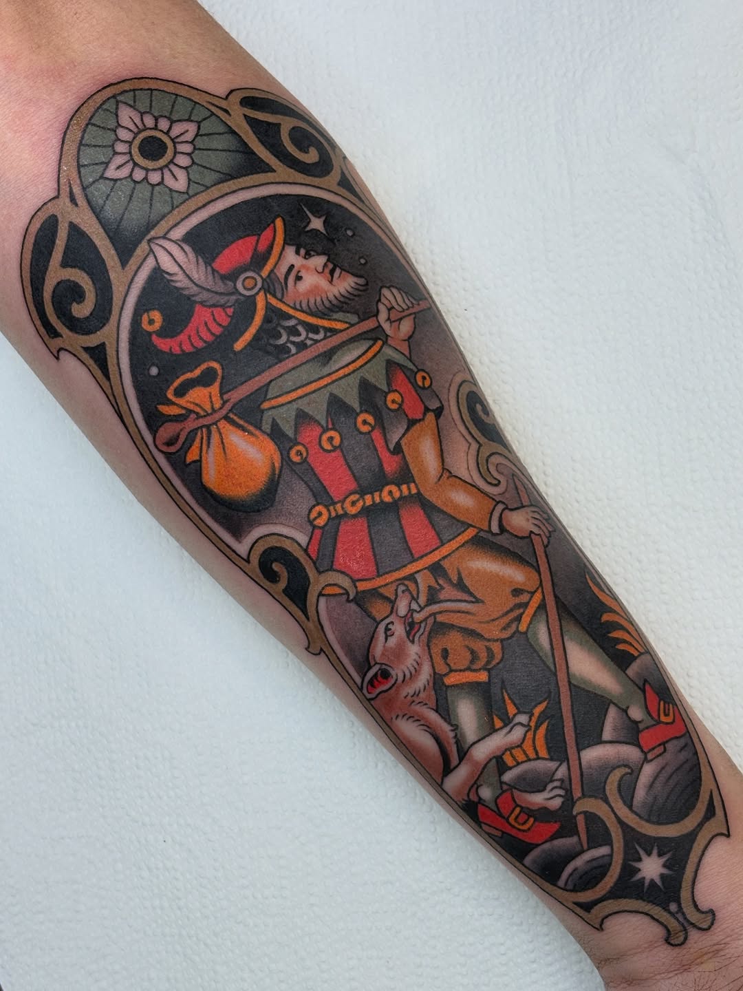 Colorful tattoo on forearm featuring a witch character with red striped dress, orange pouch, moon face, flowers, and decorative scrollwork in red, orange, and black.