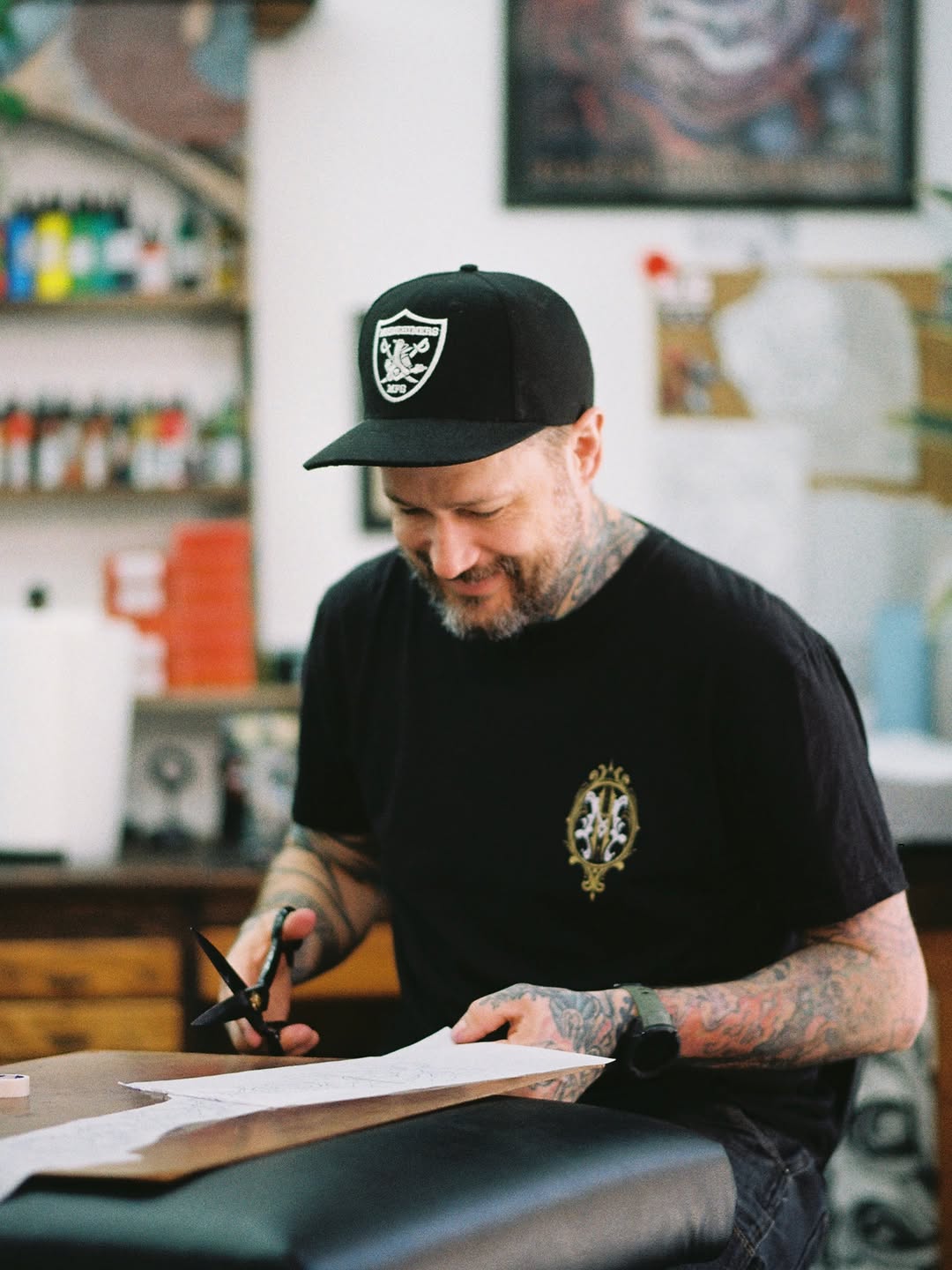 Tattoo artist with full sleeve tattoos works at a table with scissors and paper in a studio workspace.