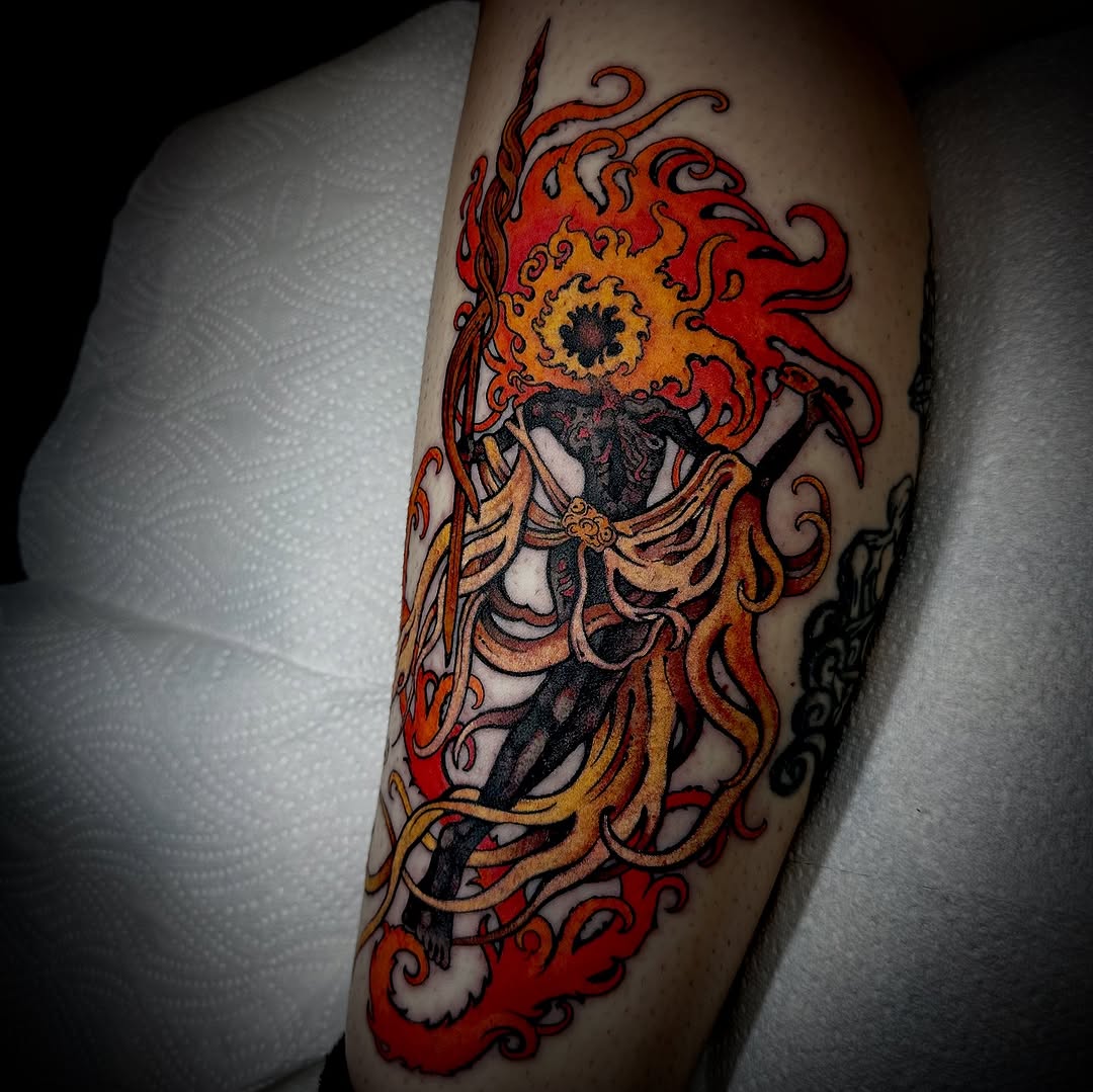 Colorful tattoo on a leg featuring a winged skeleton figure with a flaming sun head surrounded by red and orange swirling flames and decorative elements.
