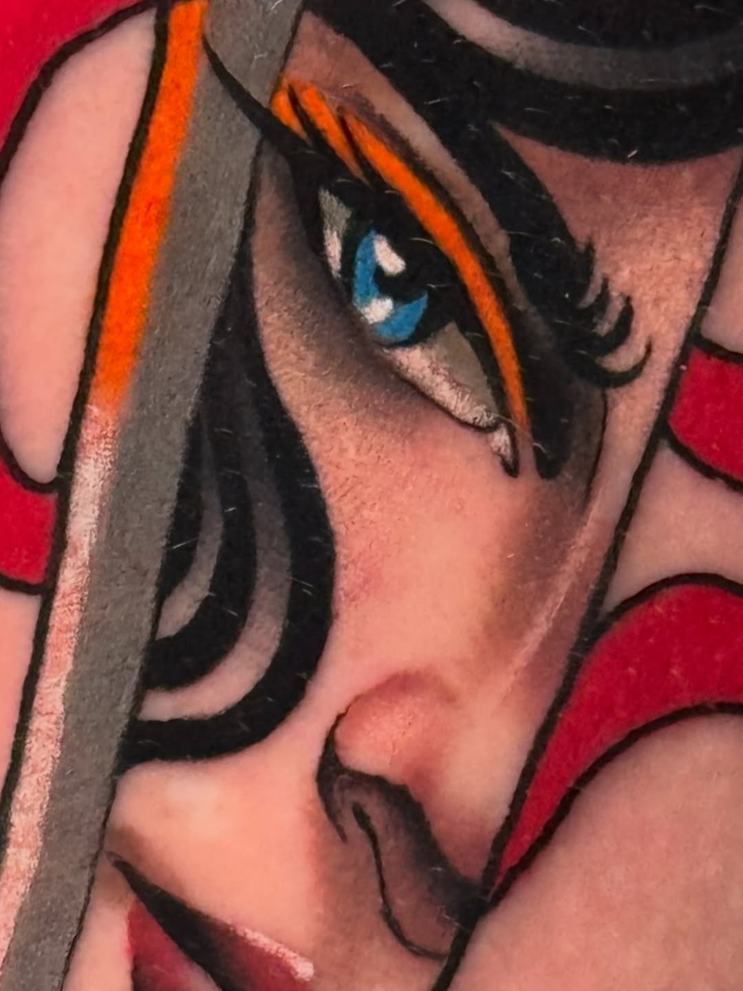 Tattoo of an anime-style female character with dark hair, blue eyes, and orange hair streaks on the upper arm against a red background.