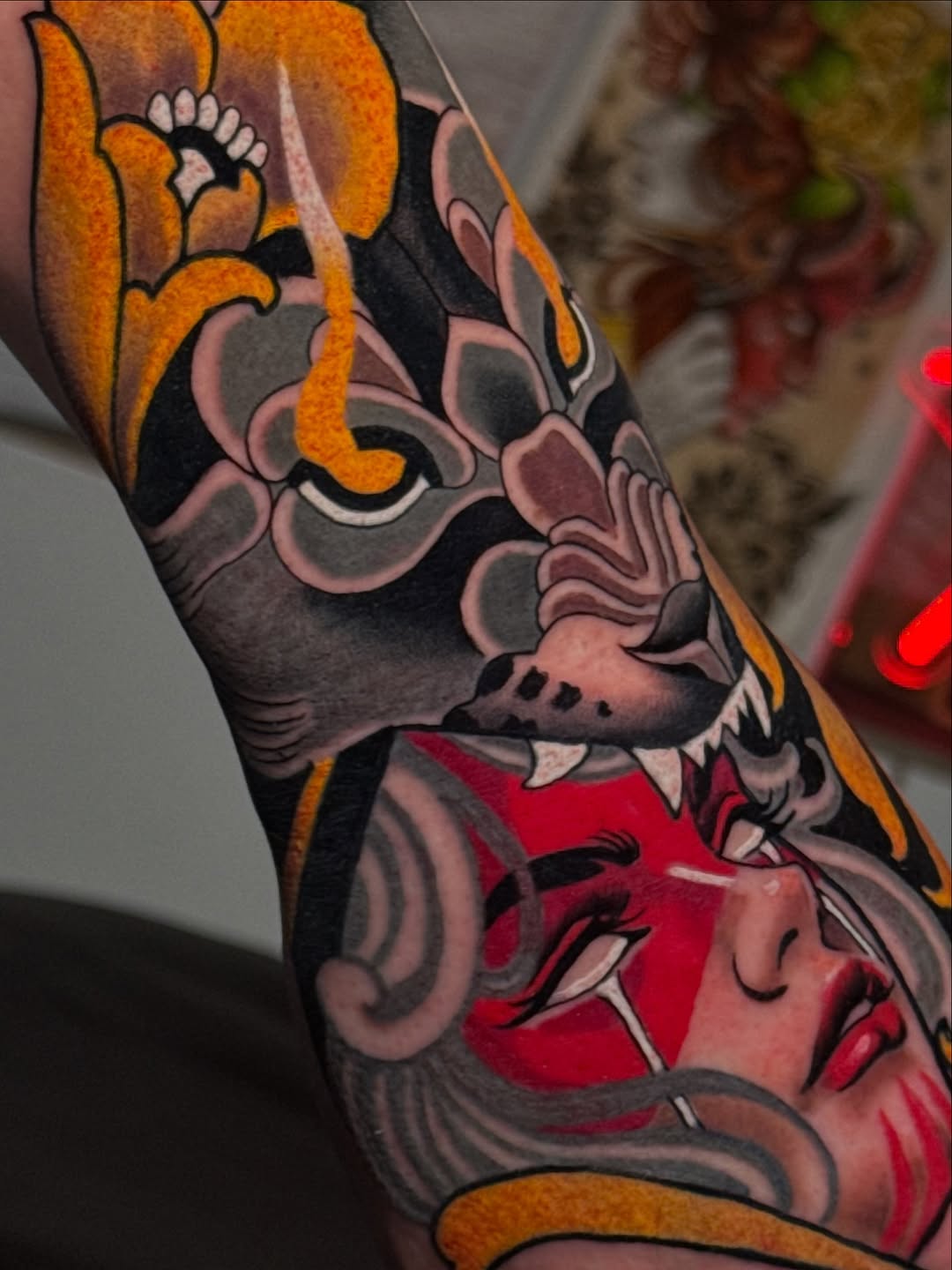 Colorful demon or oni mask tattoo on forearm featuring orange, red, black, and pink tones with intricate facial details and flowing design elements.