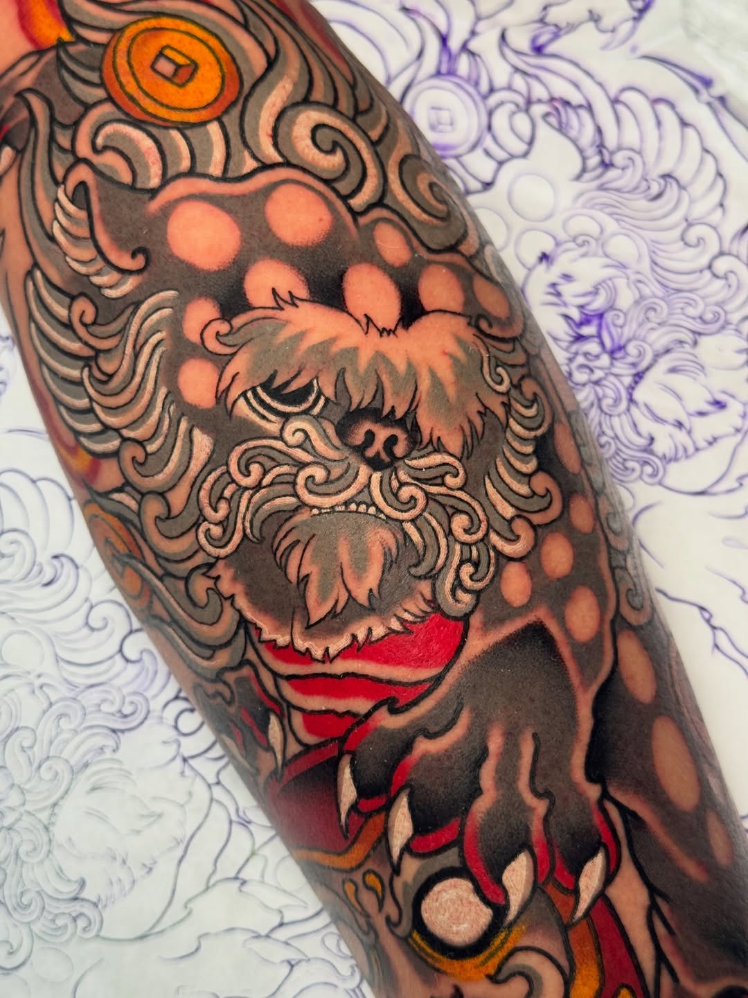 Bearded face with ornate swirling details tattooed on forearm in orange, red, black, and tan colors with decorative circular and flourish elements.