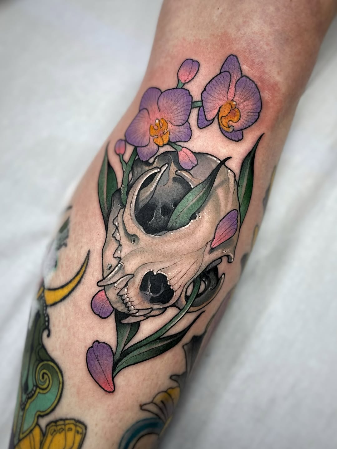Skull with purple orchids and green leaves growing from eye sockets, tattooed on forearm with yellow and teal decorative elements visible at edges.