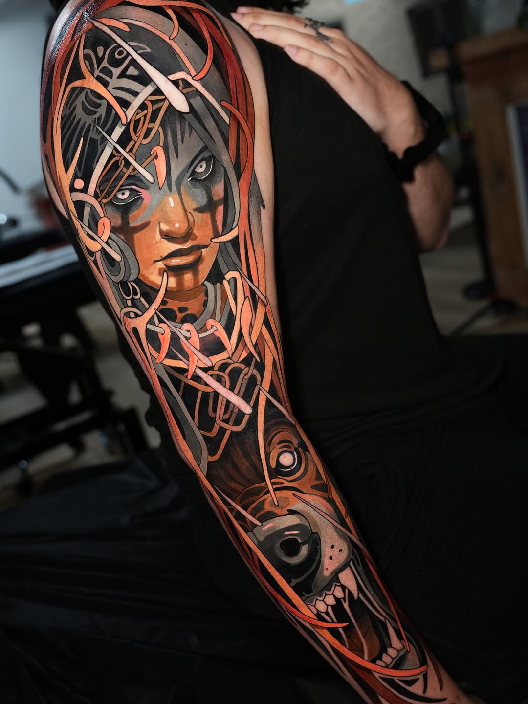 Full-sleeve arm tattoo featuring a woman's face with ornate decorative elements in orange, red, and black, with a skull design at the wrist.