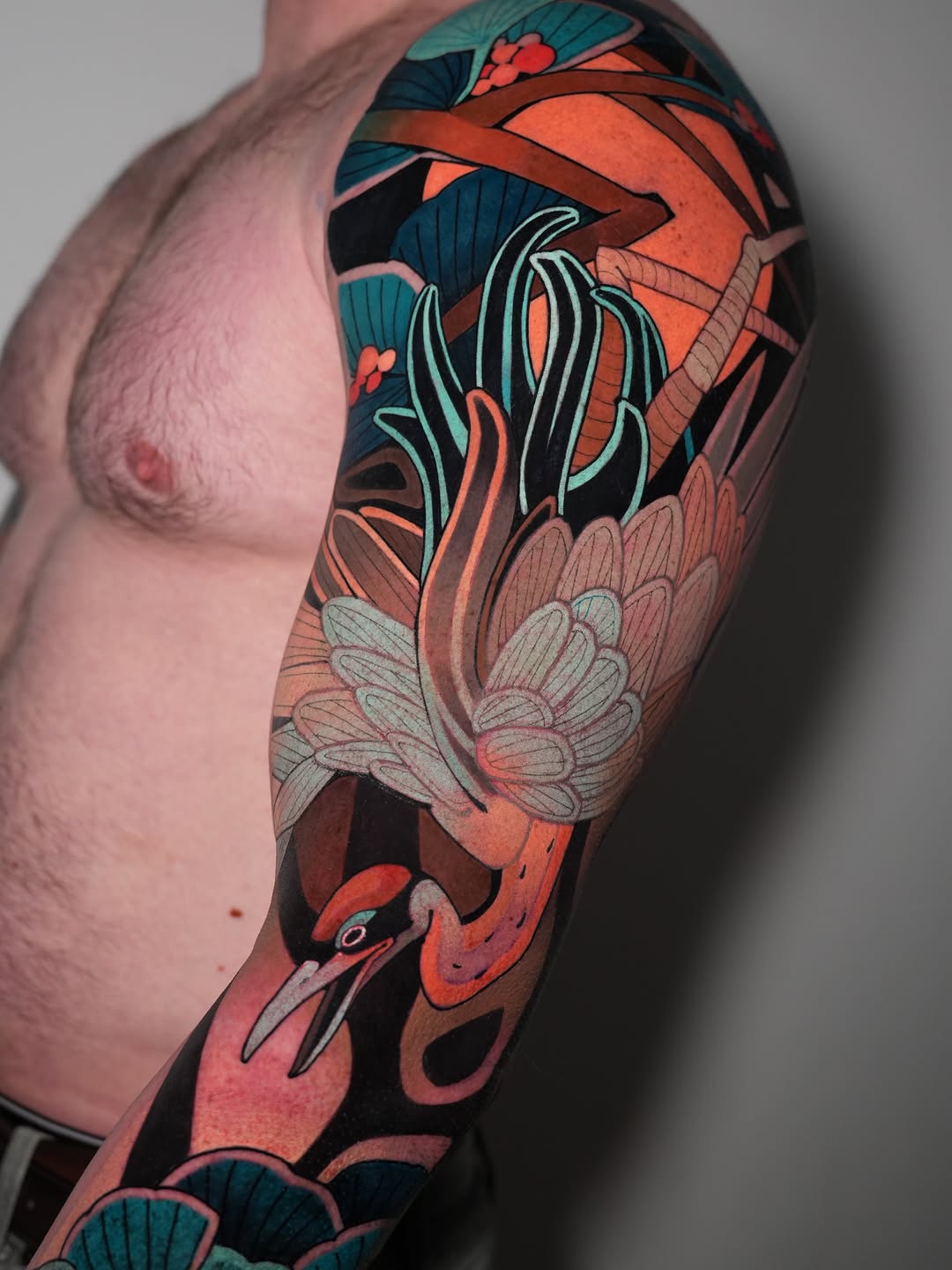 Full-color arm sleeve tattoo featuring a red bird with long teal and mint striped feathers, flowers, and fan-like foliage in coral, teal, and cream tones.