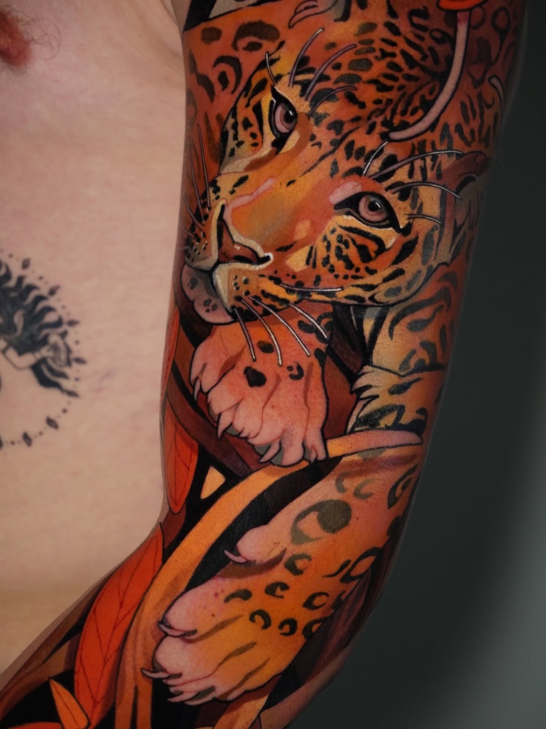 Colorful leopard head tattoo on arm featuring orange, yellow, and black tones with spotted patterns and decorative elements wrapping around the sleeve.
