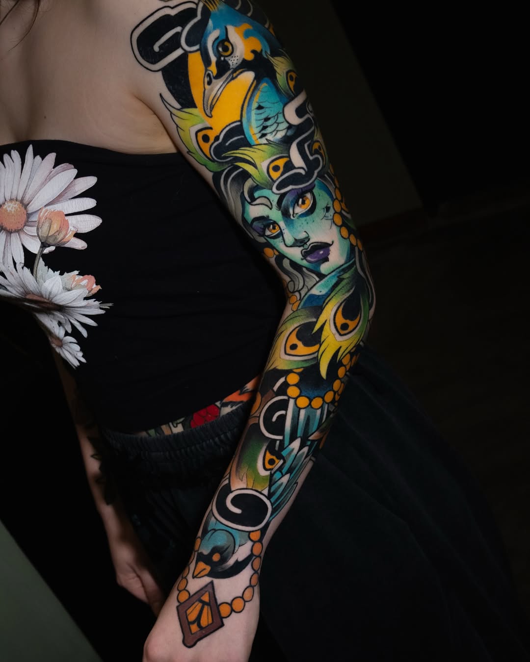 Full-sleeve arm tattoo featuring a turquoise-faced woman with yellow and black ornamental patterns, birds, and decorative elements in blue, yellow, orange, and black.