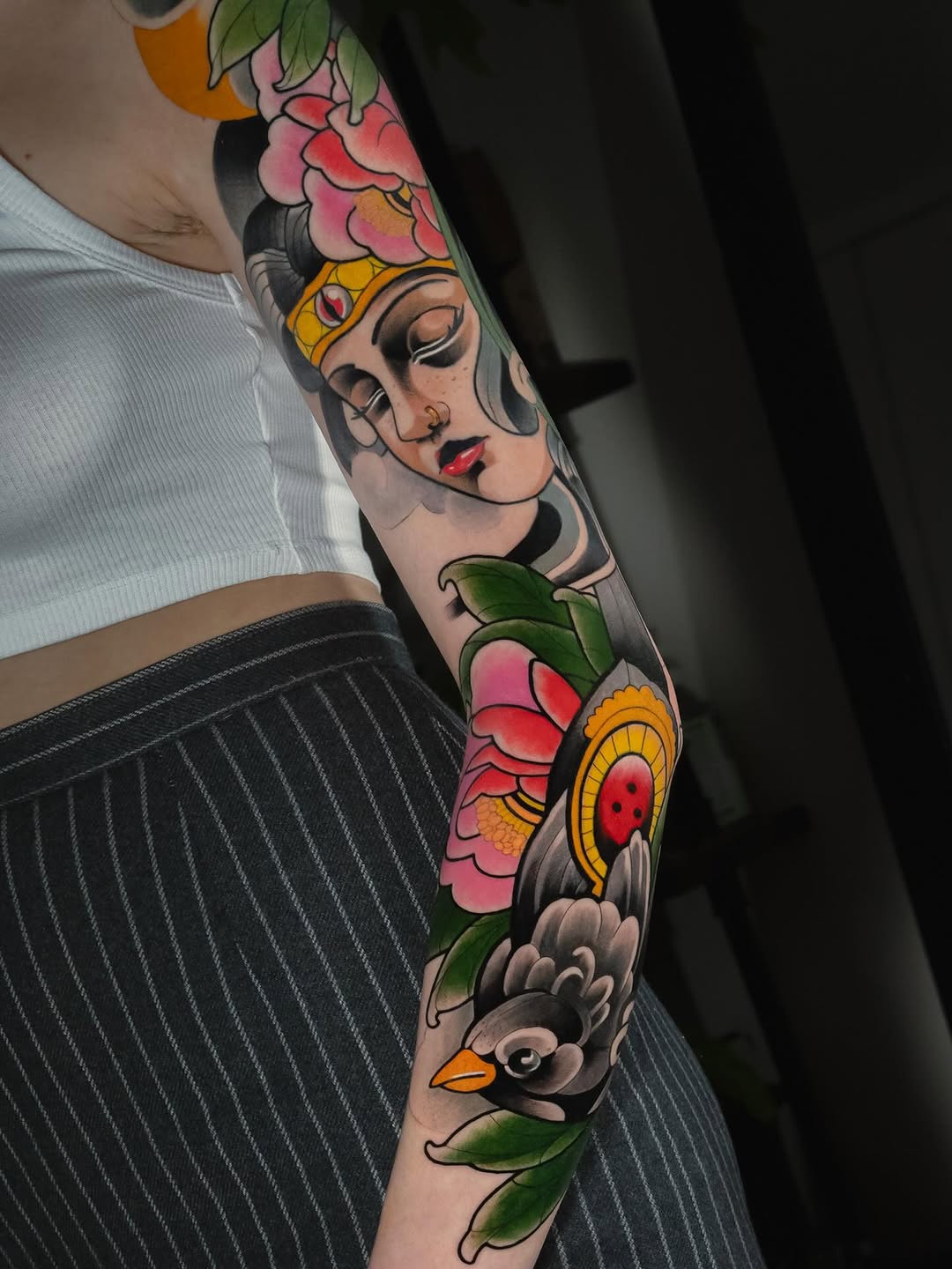 Colorful arm sleeve tattoo featuring a woman's face with a golden crown, surrounded by pink and red flowers, birds, and green leaves on skin.