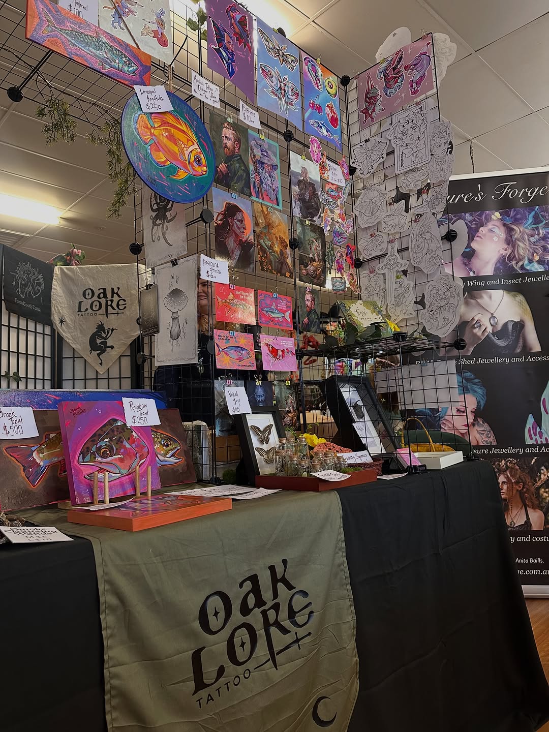 Oak Lore Tattoo studio booth displaying colorful fish, insects, and portrait artwork on walls and tables with jewelry and merchandise.