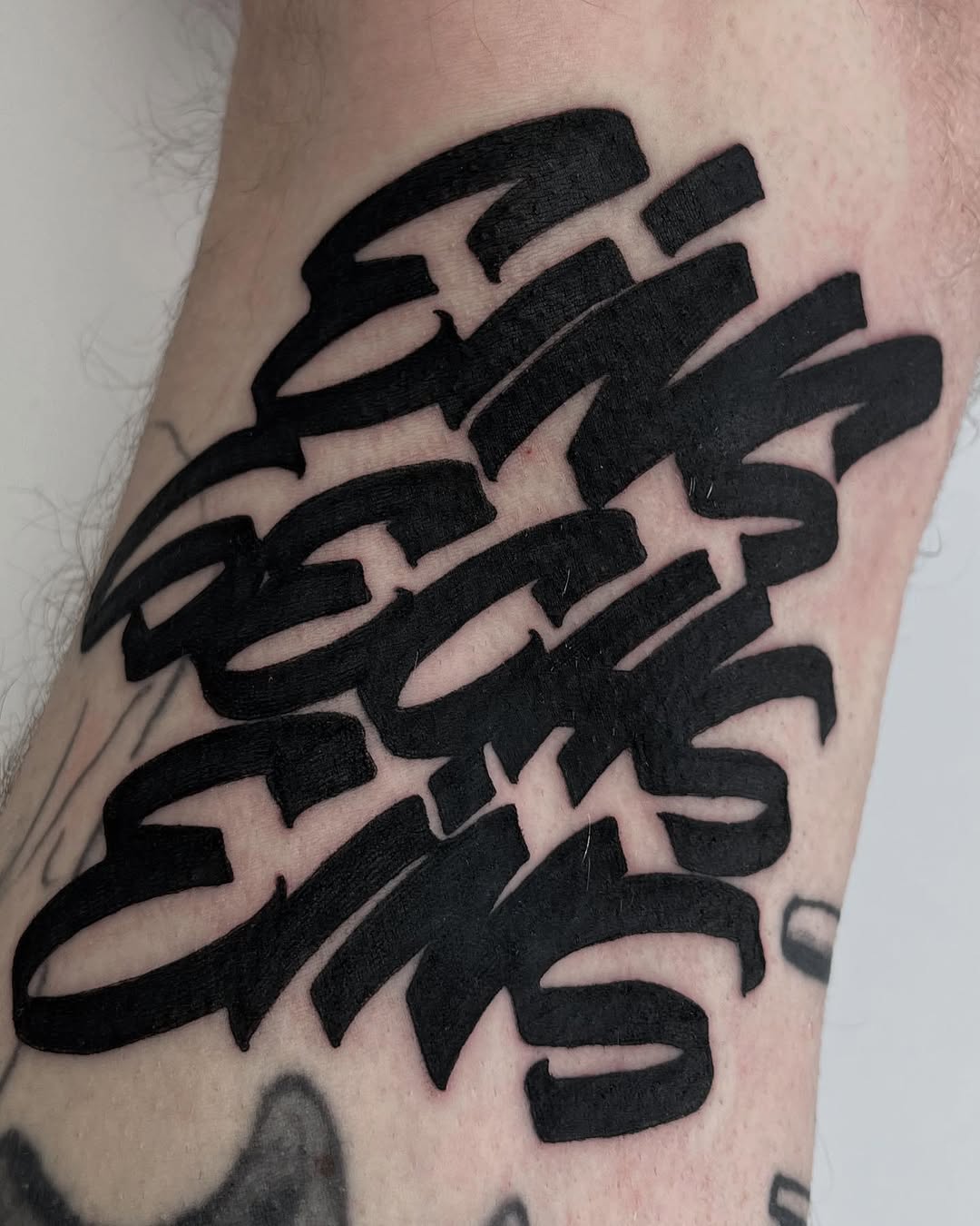 Bold black lettering tattoo with layered, dimensional text effect on the upper arm of a pale-skinned person.