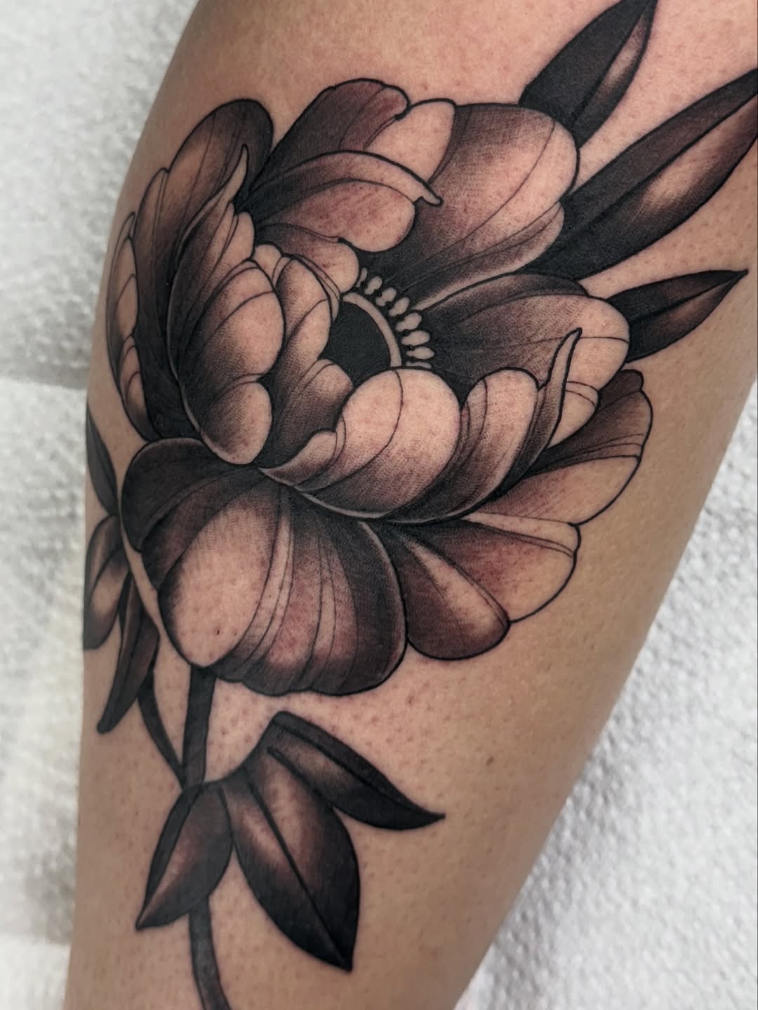 Detailed peony flower tattoo with layered petals in black and brown shading on the upper leg, accompanied by smaller blooms below.