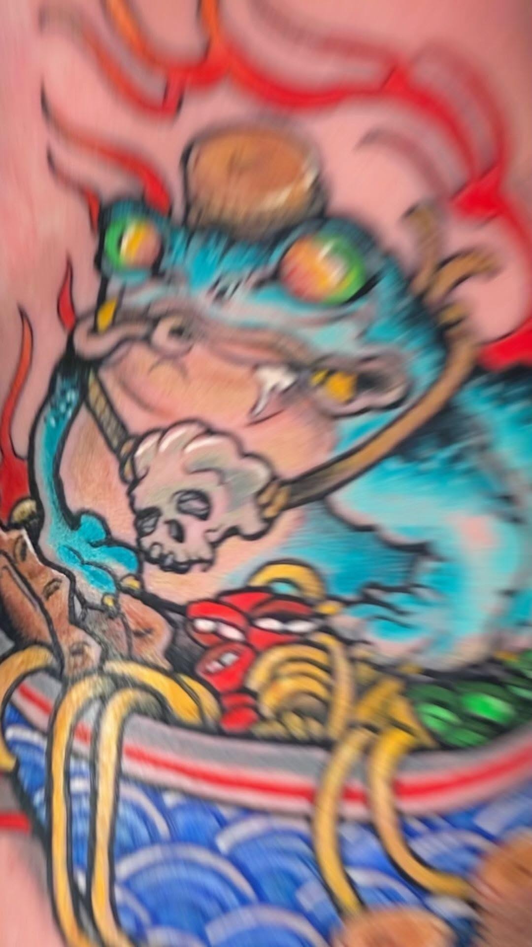 Colorful cat head tattoo with turquoise and gold accents, skull imagery, and red flame-like elements in the background on skin.