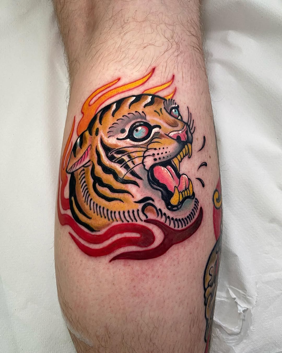 Tiger head tattoo on calf with orange and black stripes, flame details above, red wavy lines below, and a blue eye.