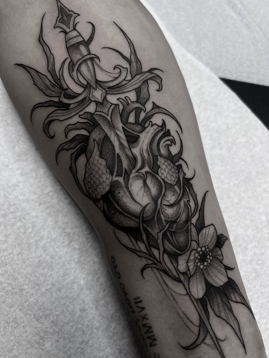 Anatomical heart with dagger, ornate scrollwork, and flowering plants in black ink on forearm, with small text visible near wrist.