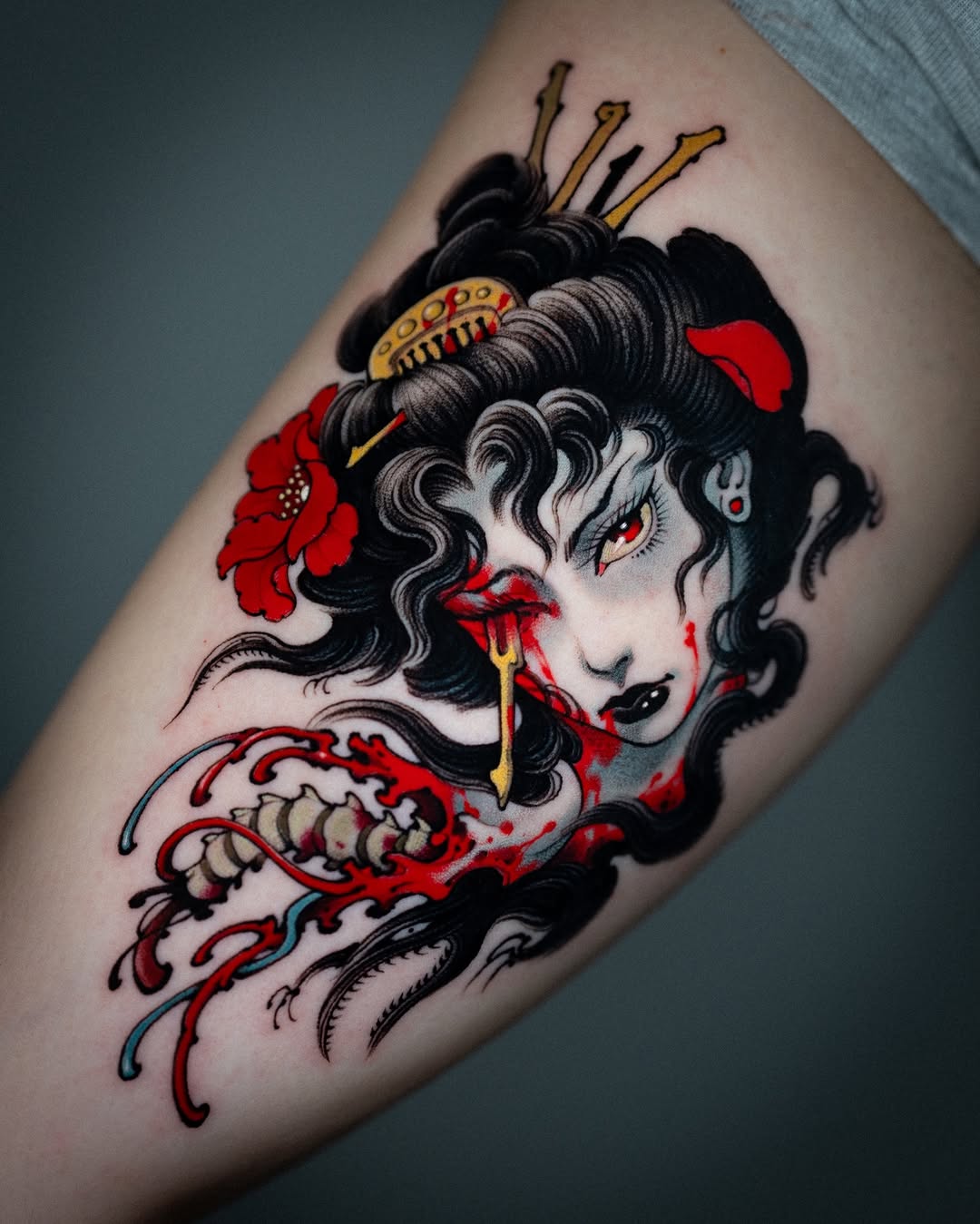 Colorful upper arm tattoo featuring a pale-faced figure with black wavy hair, crown, red flowers, and dripping red elements with gold accents.