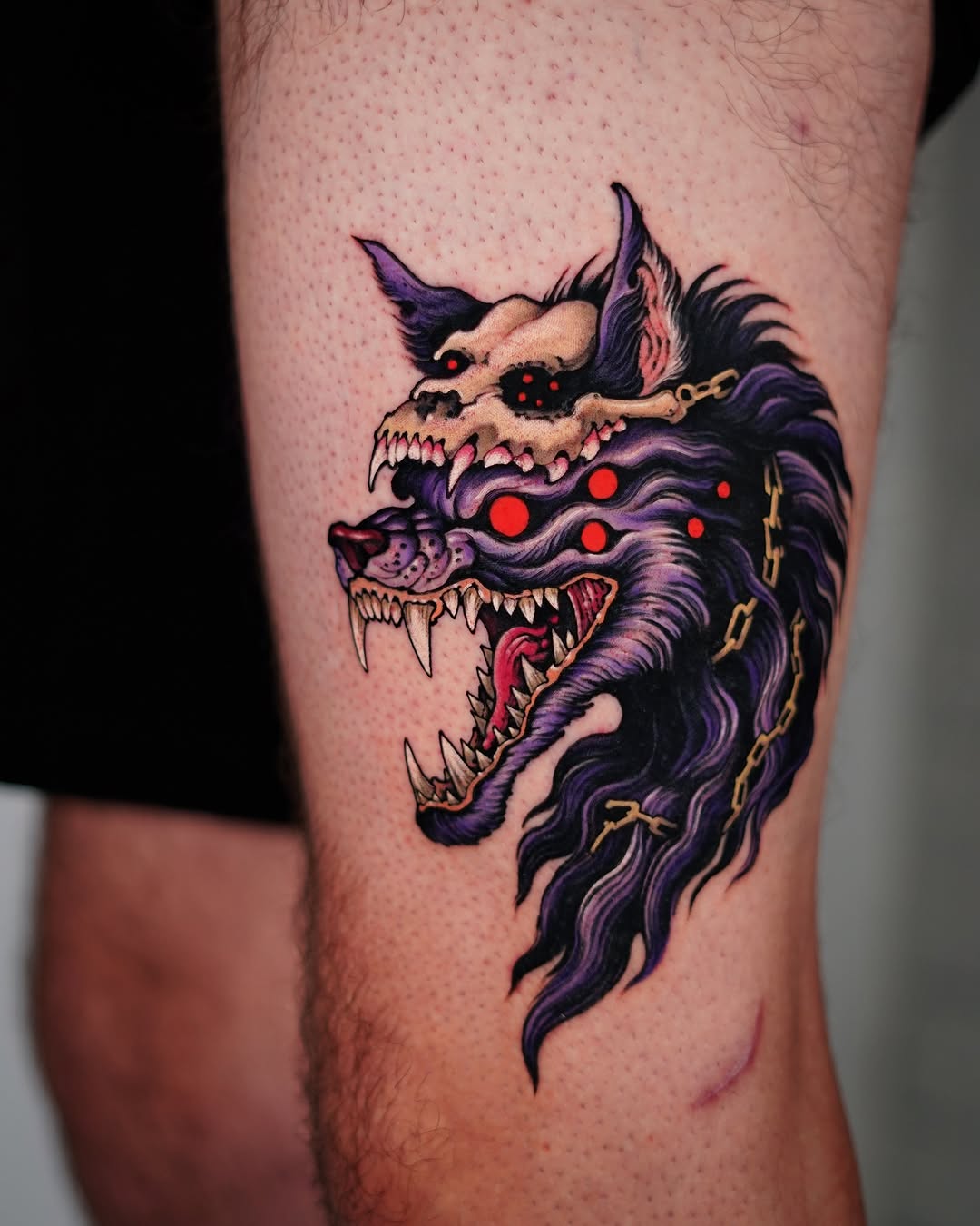 A multi-headed demon tattoo on the thigh featuring purple and black fur, skeletal faces with sharp teeth, glowing red eyes, and gold chain details.