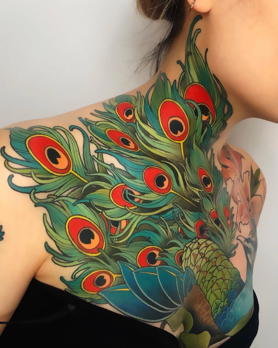 Colorful peacock feather tattoo covering the shoulder and upper back with vibrant green, red, yellow, and blue hues.