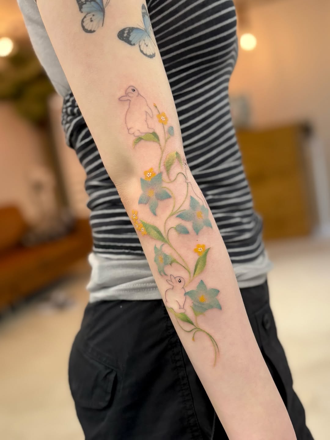 Delicate arm tattoo featuring two small birds, orange flowers with green leaves on a curved vine, and blue butterflies at the shoulder.